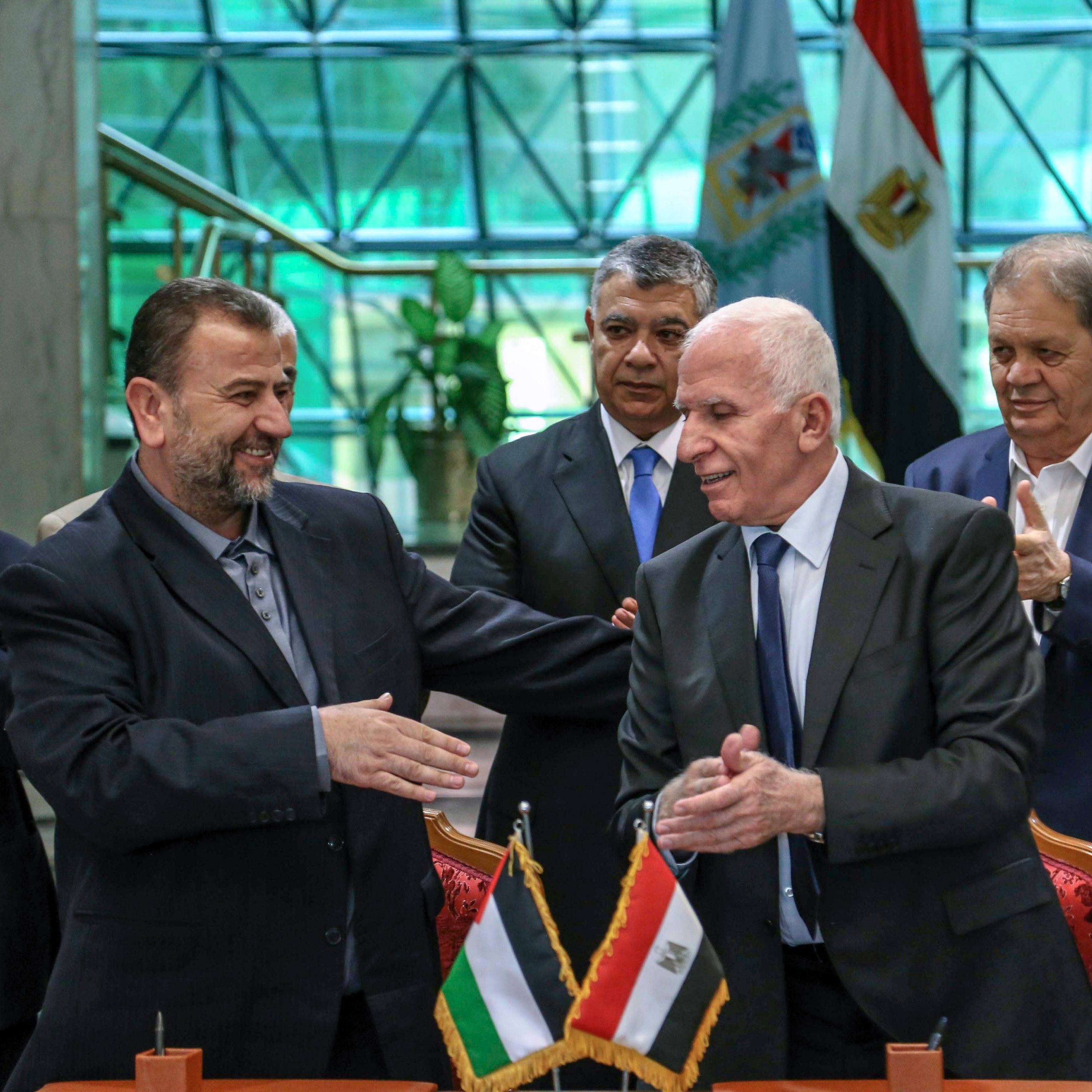 الصورة تظهر مجموعة من الشخصيات رجالية، تتواجد في مكان رسمي، حيث يتم عقد اتفاق أو توقيع. في المقدمة، يظهر شخصان shaking hands، يعبر ذلك عن اتفاق أو تعاون. الخلفية تحتوي على علمين: علم فلسطين وعلم مصر، مما يدل على موضوع النقاش أو الحدث. باقي الأشخاص في الصورة يبدون ممتنين أو يدعمون اللحظة، مما يعكس أهمية الحدث.