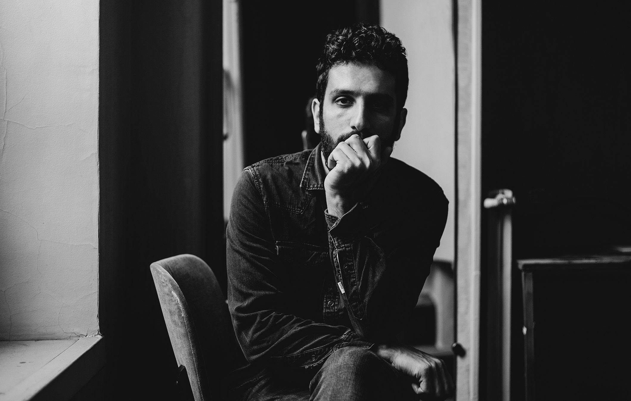 L'image montre un homme assis sur une chaise dans un intérieur sombre. Il a une barbe et porte une chemise en denim. Son expression est réfléchie, avec une main sur le menton, tandis qu'il regarde en direction de la caméra. L'éclairage met en valeur les contours de son visage et crée une ambiance introspective. Des éléments de l'environnement, comme une fenêtre et un miroir, ajoutent de la profondeur à la composition.