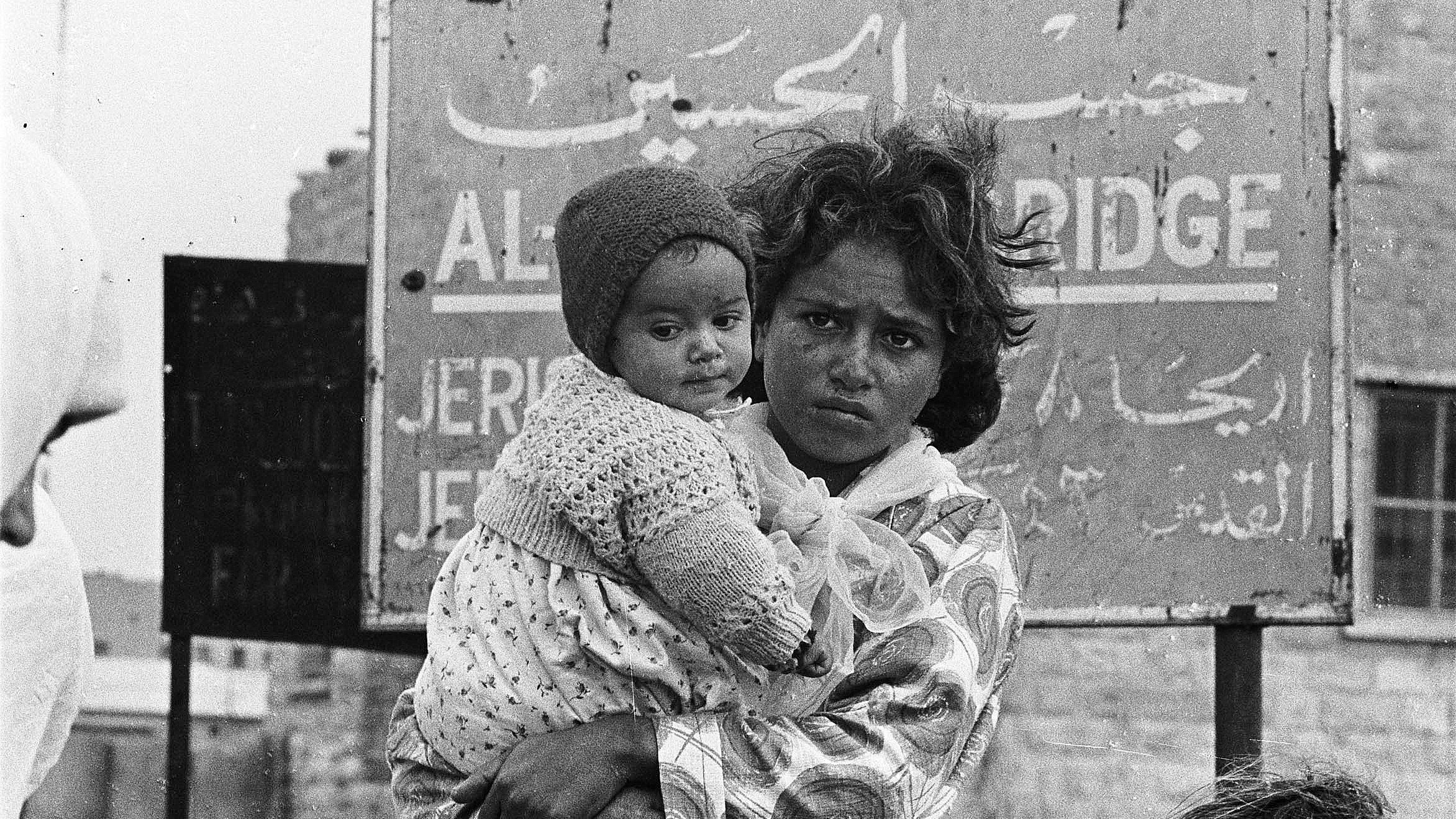 The image depicts a young woman holding a baby, both appearing to be in a challenging environment. The woman has a serious expression on her face, and her hair is unkempt. The baby, swaddled in a heavy garment, is held close to her. In the background, there's a sign written in Arabic, which seems to indicate a location, possibly related to a city or area. The overall atmosphere conveys a sense of hardship and resilience. The image is in black and white, emphasizing the emotional weight of the scene.