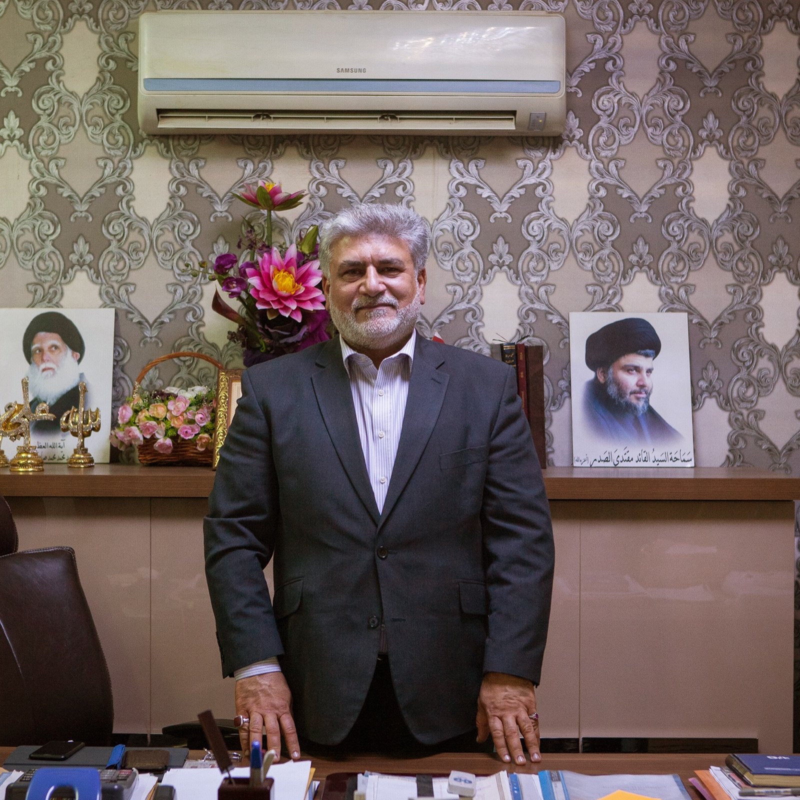 The image shows a man standing in an office setting. He is dressed in a formal suit and has a friendly expression. Behind him, there are two portraits displayed on the wall, likely of influential figures. The office has decorative elements, including a floral arrangement and a neatly organized desk with various items. The overall atmosphere conveys professionalism and authority.