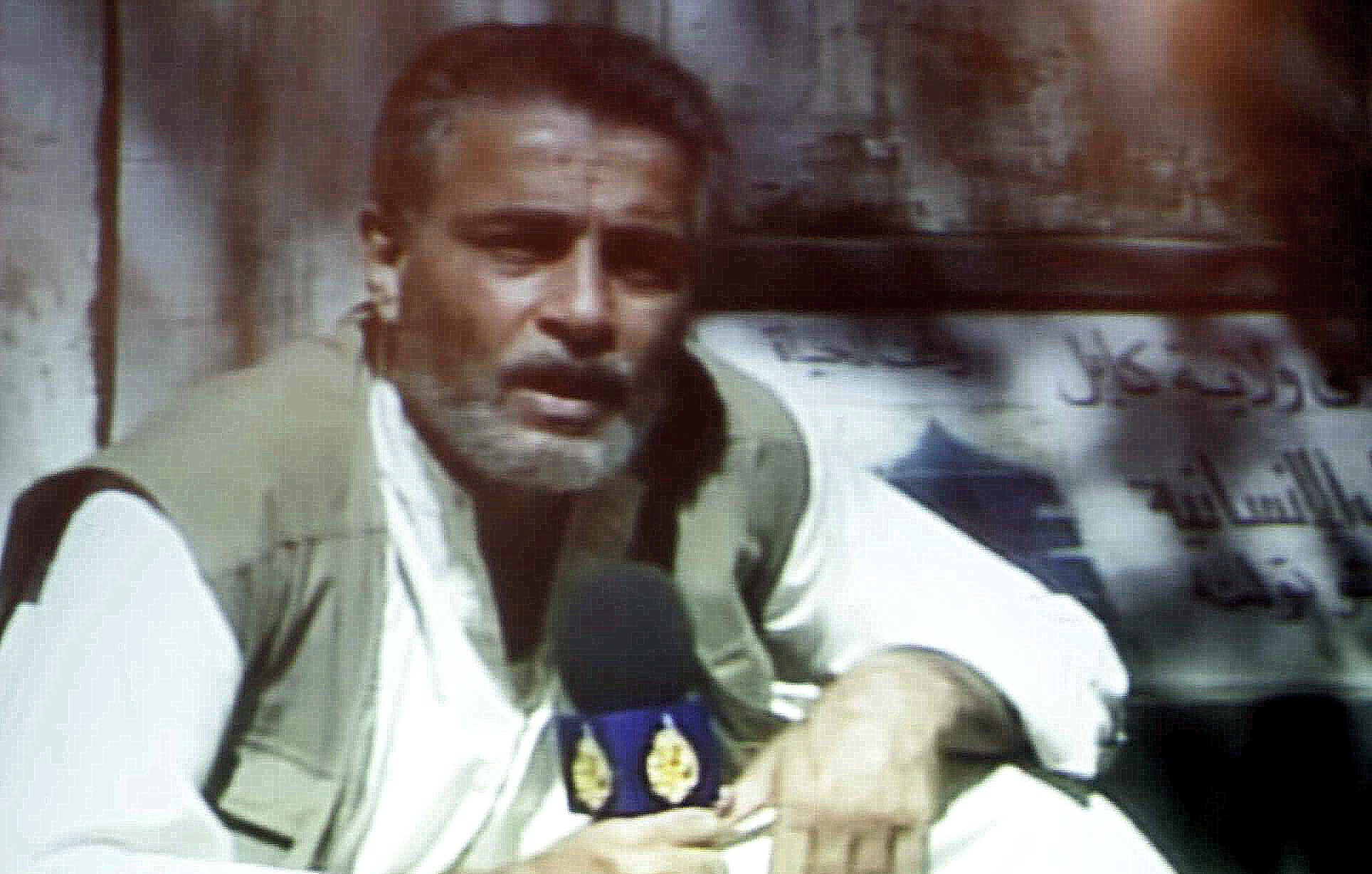 L'image montre un homme assis, tenant un micro et portant une tenue traditionnelle. Il a une barbe grise et semble parler avec intensité, probablement en train de faire un reportage. En arrière-plan, il y a un panneau ou une affiche qui n'est pas clairement visible. L'éclairage de l'image semble un peu flou, ajoutant une ambiance particulière à la scène.