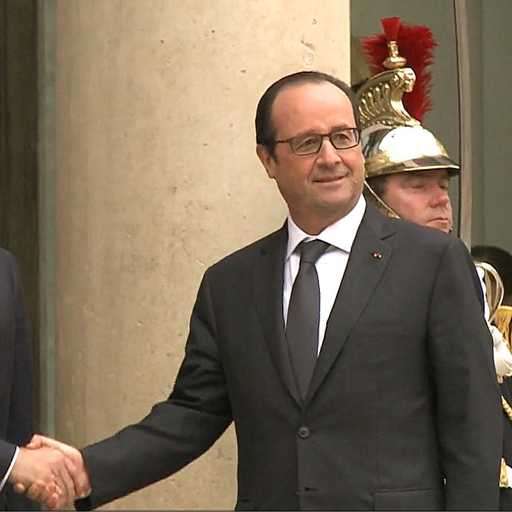 L'image montre deux hommes en costume, serrant la main l'un de l'autre. L'un est à gauche et semble avoir une expression amicale, tandis que l'autre se trouve à droite, affichant également un sourire. En arrière-plan, on aperçoit un soldat en uniforme. L'environnement semble être formel, probablement devant un bâtiment gouvernemental.