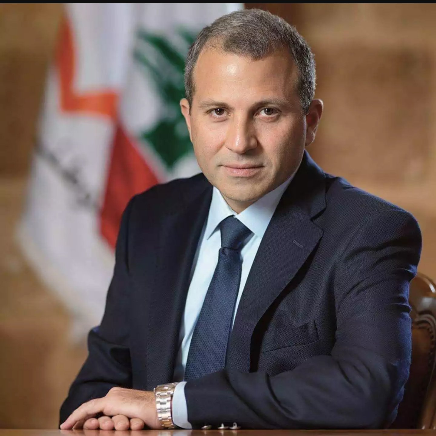 الصورة تظهر رجلًا يرتدي بدلة رسمية داكنة، مع قميص أبيض وروابط عنق زرقاء. يجلس بشكل مشي هادئ وملامحه تظهر الثقة. في الخلفية، تتواجد علم لبنان، مما يشير إلى احتمال ارتباطه بالسياسة أو الحكومة في لبنان. الجو العام للصورة يعكس الرسمية والجدية.