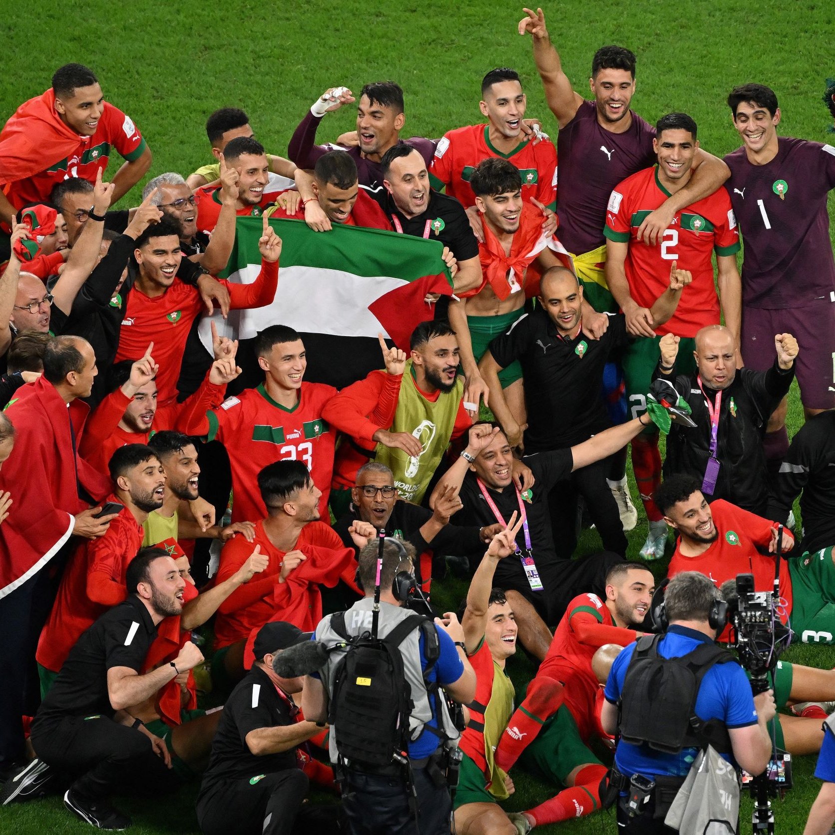 L'immagine mostra un gruppo di giocatori di calcio e membri dello staff, tutti gioiosi e festanti, mentre celebrano una vittoria. Sono vestiti con magliette rosse e verdi, i colori della nazionale marocchina, e alcuni di loro tengono in alto una bandiera. Il gruppo è radunato attorno a un portiere in uniforme, e l'atmosfera è di esultanza e festa. I volti sorridenti e le pose celebrative trasmettono un forte senso di trionfo e unità.
