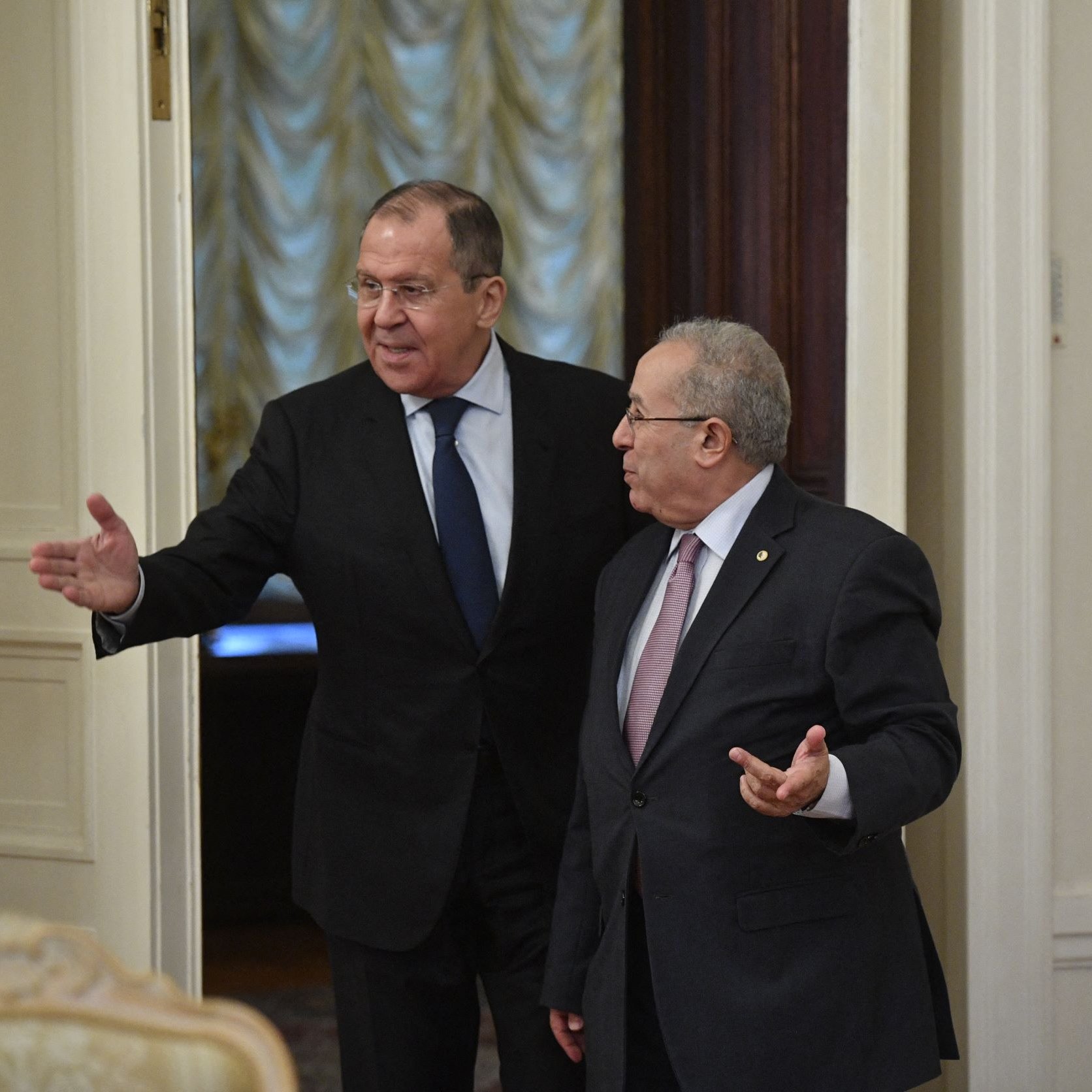 این تصویر دو مرد را نشان می‌دهد که در یک فضای داخلی ملاقات می‌کنند. یکی از آنها لباس رسمی با کت و شلوار به تن دارد و به سمت دوربین اشاره می‌کند. دیگری نیز در حالی که لباسی رسمی به تن دارد، به طرف مقابل نگاه می‌کند و دستش را به نشانه صحبت دراز کرده است. محیط اطراف شامل دیوارهایی با طراحی کلاسیک و پرده‌هایی به رنگ ملایم است. به نظر می‌رسد این ملاقات در یک مکان رسمی انجام می‌شود.