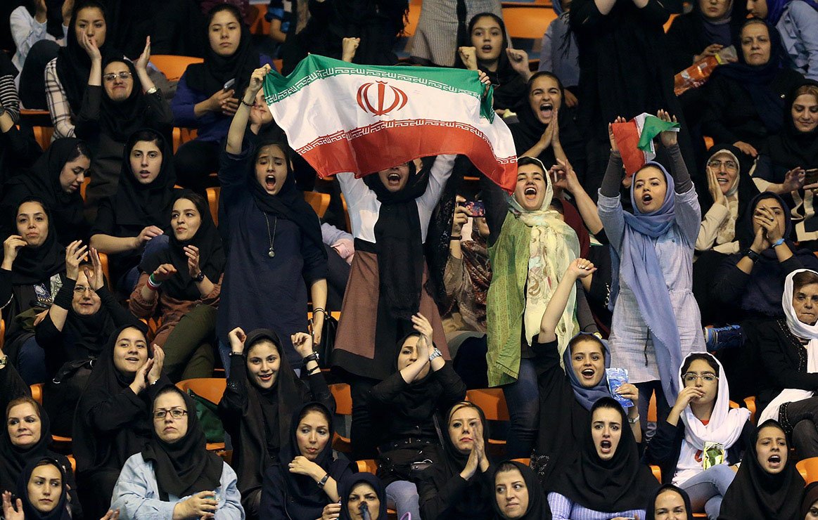 L'image montre un groupe de femmes dans une arène, probablement lors d'un événement sportif. Elles portent des vêtements traditionnels et modernes, et certaines d'entre elles agitent un drapeau iranien, exprimant leur soutien et leur enthousiasme. Les visages sont joyeux et animés, et l'atmosphère semble festive. Ce moment illustre la participation et la passion des femmes pour le sport en Iran.