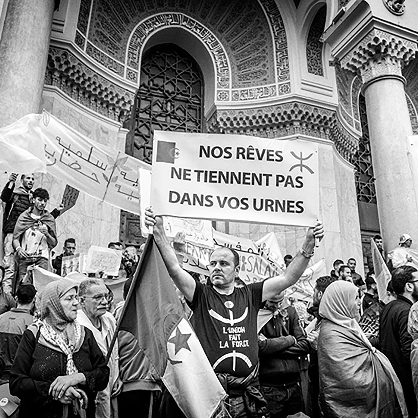 تظهر الصورة مجموعة من المتظاهرين في ساحة عامة، حيث يحمل أحدهم لافتة مكتوبًا عليها باللغة الفرنسية "أحلامنا لا تتواجد في صناديق الاقتراع". يُظهر المشهد حشدًا من الناس يتجمعون معًا، مع تباين واضح بين الأعمار والملابس. بعض الأشخاص يرتدون الأوشحة أو الحجاب، مما يعكس التنوع الثقافي. تعكس الأجواء إحساسًا قويًا بالتضامن والمشاركة في قضية معينة.