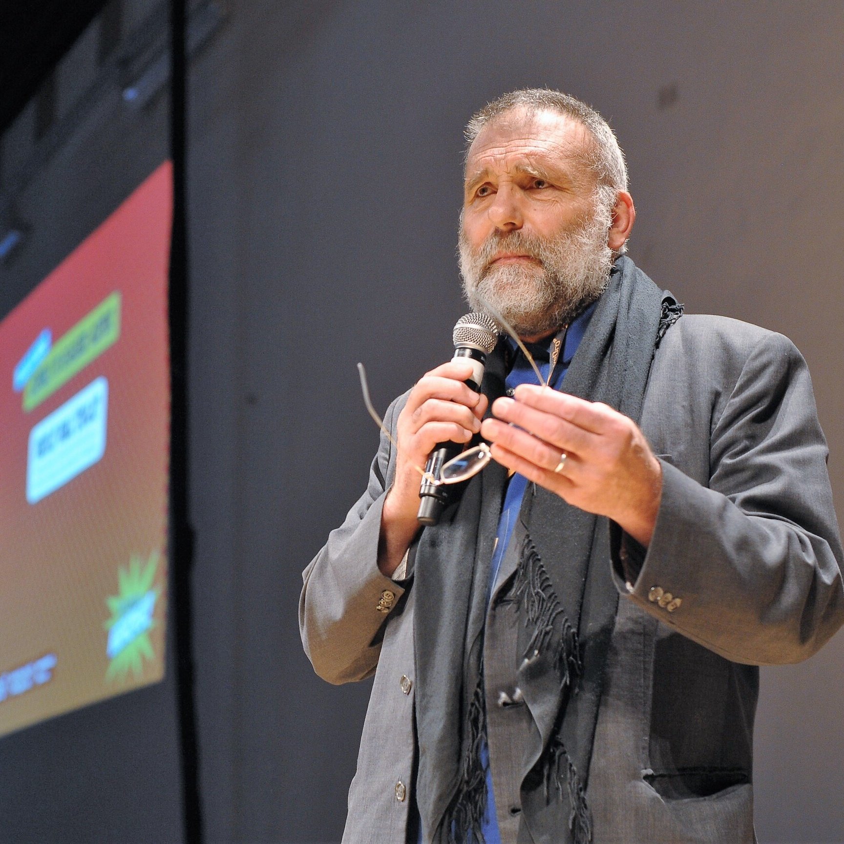 Nell'immagine si vede un uomo, probabilmente un relatore, che sta parlando al pubblico. Indossa un abito elegante e ha una sciarpa scura. Tiene un microfono in mano e sembra essere coinvolto in un discorso. Sullo sfondo c'è un grande schermo con delle grafiche colorate, ma il contenuto di ciò che viene mostrato non è visibile. L'atmosfera dell'immagine sembra essere formale e interattiva.