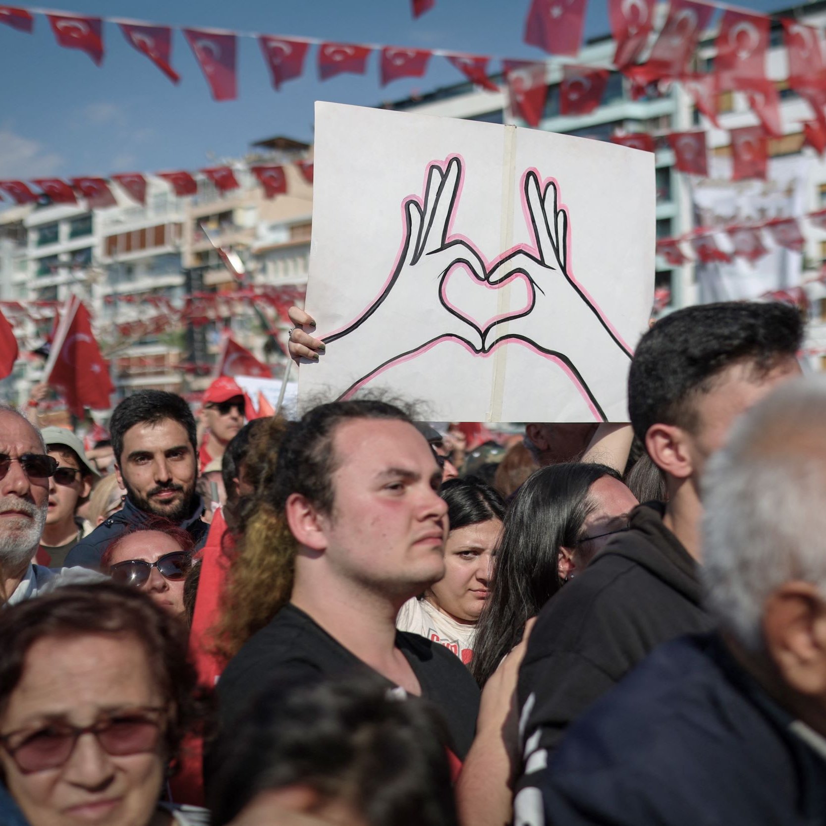 La imagen muestra una multitud reunida en un evento, posiblemente una manifestación o celebración. En primer plano, hay un hombre que sostiene un cartel en forma de corazón, dibujado con las manos. La multitud parece diversa y está vestida con ropa casual. Al fondo, hay banderas rojas con la insignia de Turquía, lo que sugiere un contexto nacional. La atmósfera parece ser de unidad y orgullo colectivo.
