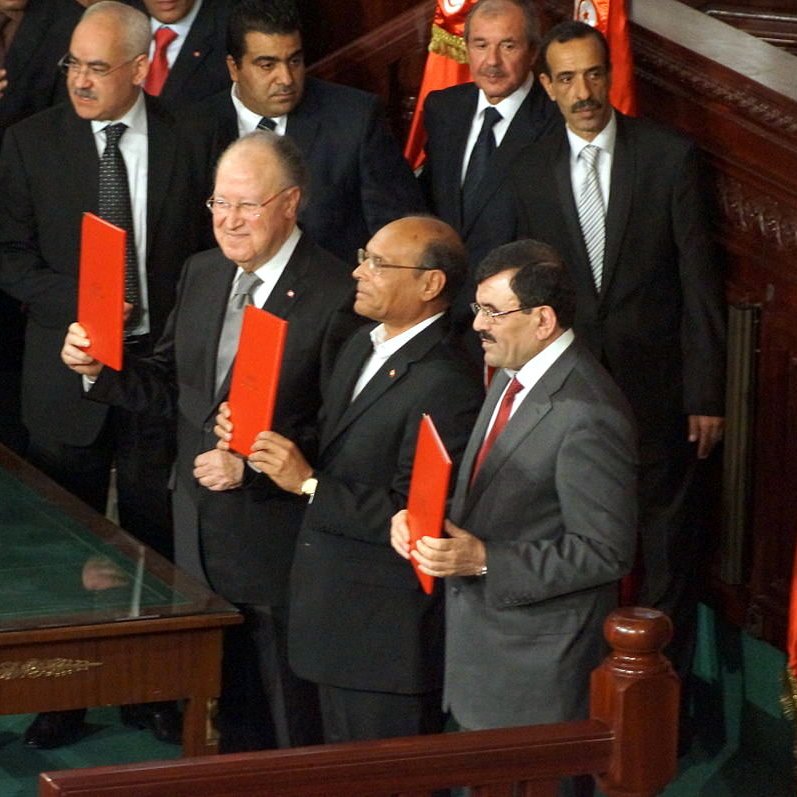 L'image montre un groupe d'hommes en costume, apparemment dans un cadre officiel, probablement lors d'une cérémonie ou d'un événement politique. Ils tiennent des documents en main, et l'ambiance semble solennelle. On peut voir une table en bois devant eux ainsi que des drapeaux en arrière-plan. Les personnes présentes semblent applaudir ou se concentrer sur l'événement.