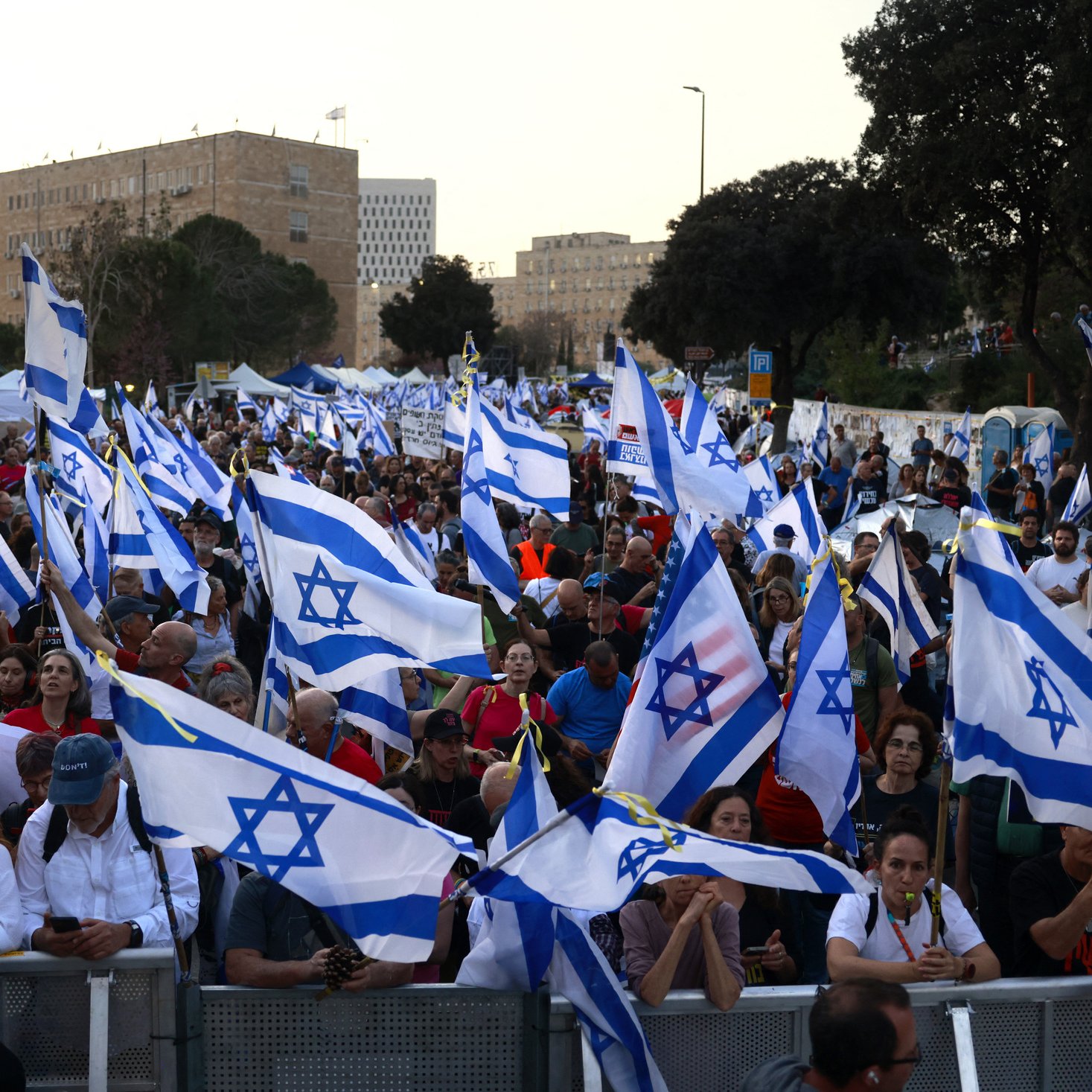 La imagen muestra una multitud de personas en una manifestación, ondeando banderas de Israel. La escena parece vibrante y llena de energía, con una gran cantidad de asistentes que levantan las banderas en un ambiente de protesta o celebración. En el fondo se pueden apreciar árboles y estructuras, lo que sugiere que el evento tiene lugar al aire libre, probablemente en una ciudad. La multitud parece diversa y está unida por una causa común.