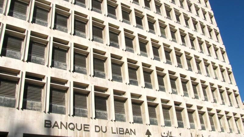 الصورة تظهر واجهة مبنى كبير يتكون من عدة طوابق، ويتميز بتصميم معماري حديث وبسيط. الجدران مصنوعة من الحجر أو الخرسانة، مع نوافذ موزعة بشكل منتظم. يظهر اسم البنك "BANQUE DU LIBAN" باللغة الفرنسية، مع إضافة عبارة باللغة العربية "مـصرف لبنان" تحتها. المبنى يعكس الطابع المؤسسي ويبدو أنه يقع تحت ضوء الشمس، مما يعطي شعوراً بالوضوح والنقاء.