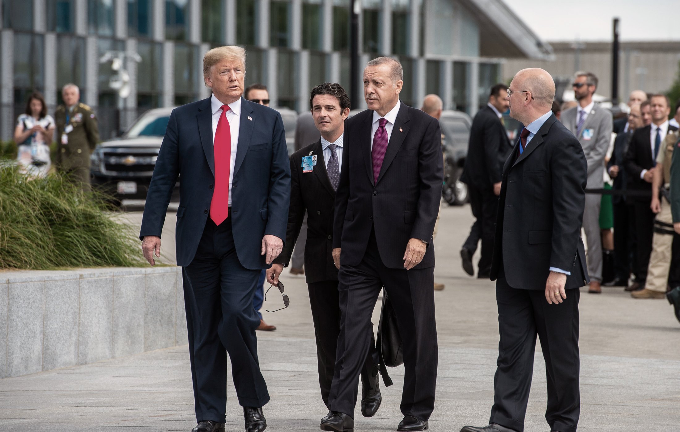 L'image montre un groupe d'hommes en costumes, apparemment dans un cadre formel, probablement lors d'un événement diplomatique. Deux hommes, respectivement en costume noir et rouge, marchent côte à côte, tandis que d'autres les suivent. L'arrière-plan indique un environnement urbain moderne, avec des véhicules et des bâtiments. L'atmosphère semble sérieuse, éventuellement liée à des discussions ou des négociations importantes.