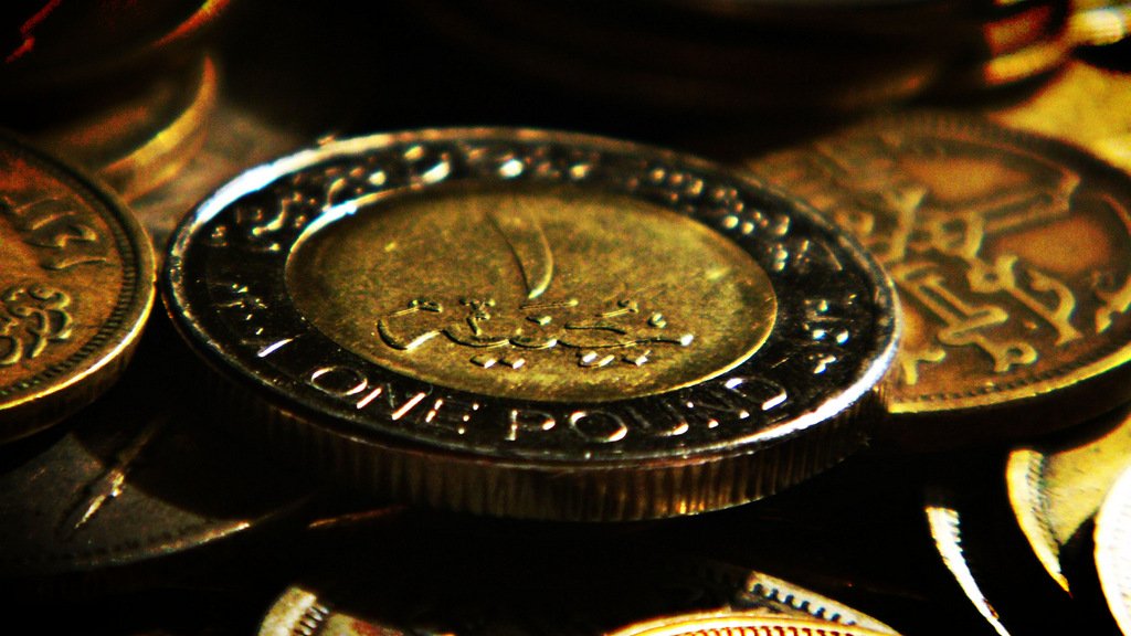 L'image montre plusieurs pièces de monnaie, avec une pièce de un pound en avant-plan. La pièce est en métal, et on peut voir des inscriptions en arabe sur sa surface, ainsi que les mots "ONE POUND". L'arrière-plan est flou, composé de plusieurs autres pièces de monnaie qui ajoutent à la richesse visuelle de la composition. Les pièces semblent avoir un éclat doré, ce qui attire l'attention sur le sujet principal.