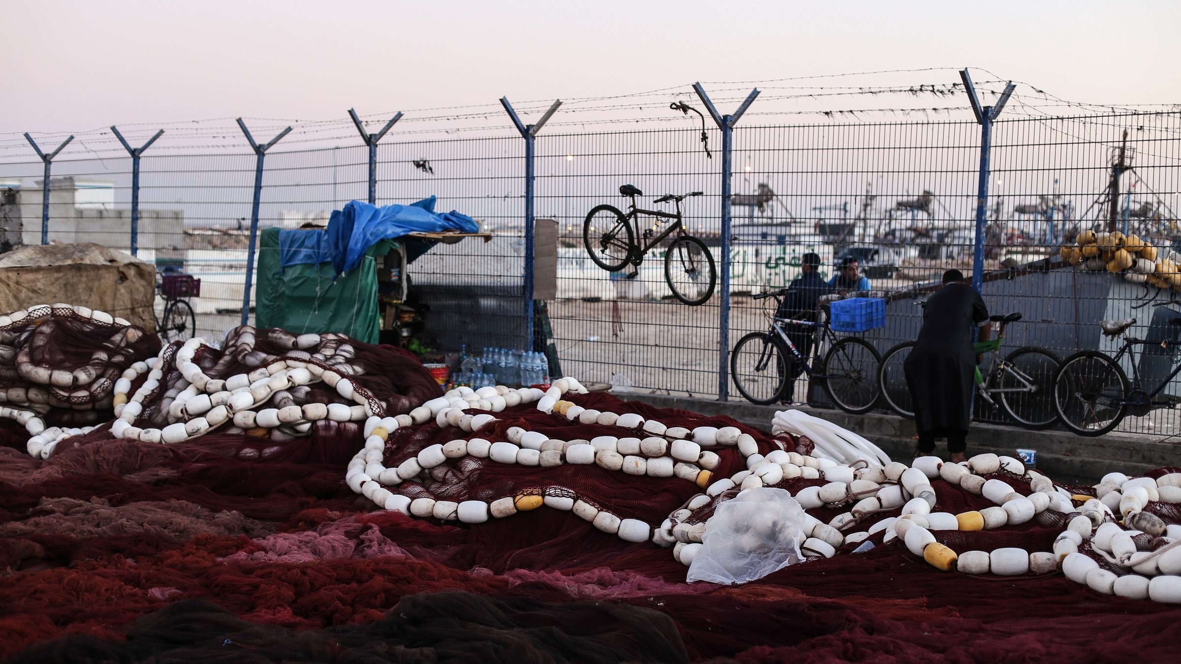 L'image montre un environnement portuaire où se trouvent des filets de pêche. De grandes cordes blanches, probablement des bouées, sont éparpillées sur le sol, mêlées à des filets de couleur rouge foncé. En arrière-plan, on aperçoit une clôture barbelée qui délimite l'espace, ainsi que quelques bicyclettes accrochées. On peut aussi voir des personnes se déplacer, l'une d'elles étant vêtue d'une tenue traditionnelle. Le ciel est légèrement teinté, indiquant peut-être le début de la soirée. L'atmosphère semble calme, mais il y a une impression d'activité liée à la pêche.