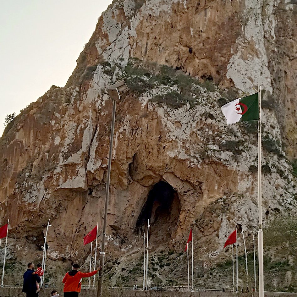 La imagen muestra un paisaje montañoso con una gran roca o acantilado en el centro, que tiene una cueva visible en su base. A su alrededor, hay varias banderas ondeando, entre las cuales algunas son de Argelia, ya que tienen un diseño distintivo. En la parte inferior de la imagen, se pueden ver algunas personas caminando y disfrutando del lugar. El cielo tiene un tono claro, posiblemente al atardecer. La escena refleja un ambiente natural y un sitio de interés cultural o histórico.