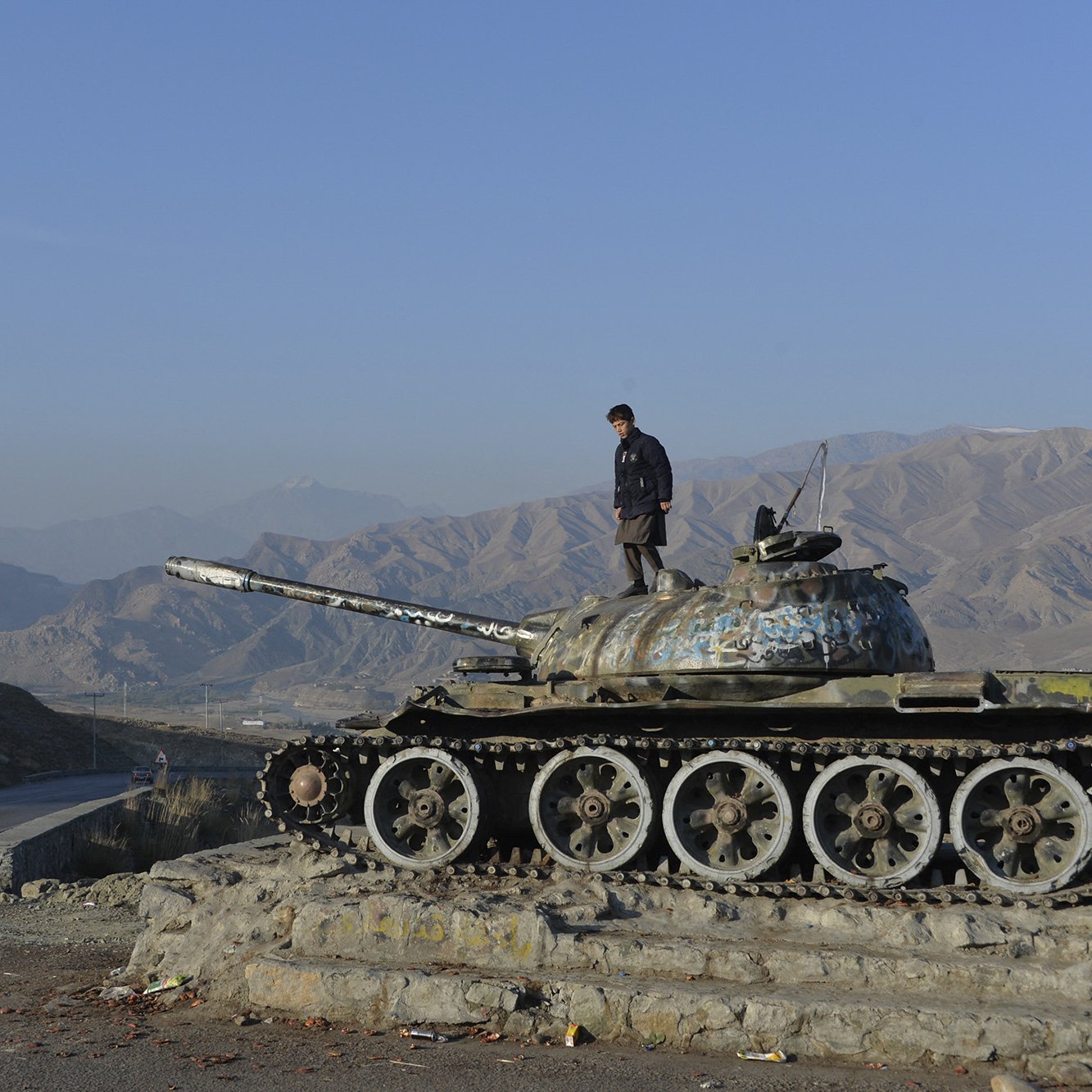 The image depicts a person standing atop an old tank, which is positioned on a small rise alongside a road. The tank appears weathered and has a layer of rust. In the background, there are rolling mountains under a clear blue sky, creating a stark contrast with the tank's dilapidated condition. A car can be seen on the road nearby, suggesting a sense of movement in this otherwise desolate landscape. Overall, the scene conveys themes of history and the passage of time in a rugged environment.