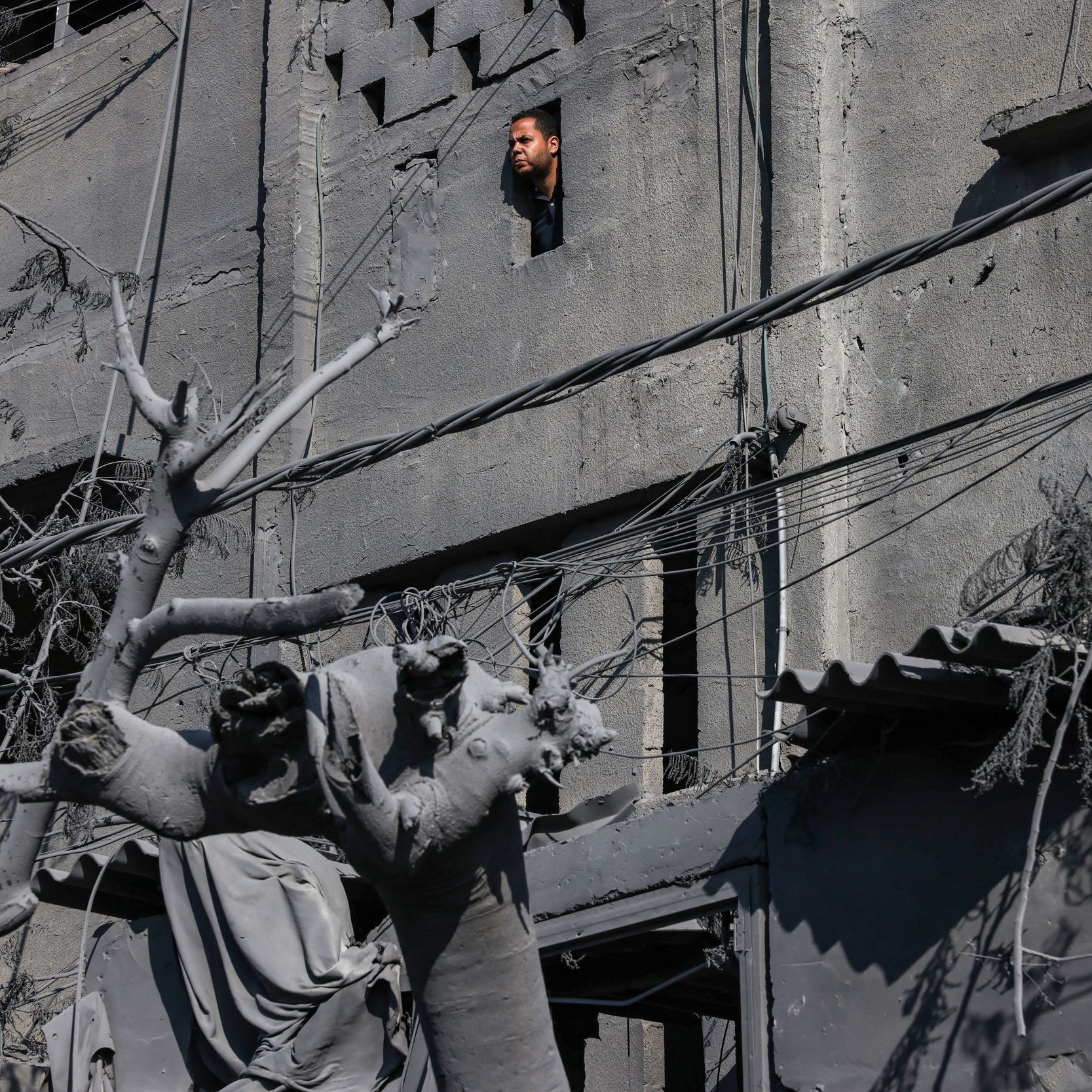 L'image montre un bâtiment en ruines avec des murs de béton gris, où l'on aperçoit une fenêtre. À travers cette fenêtre, une personne regarde vers l'extérieur. Au premier plan, on peut voir des débris et une statue partiellement détruite, entourée de fils électriques et de restes de végétation, ce qui donne une impression de désolation. L'environnement semble touché par un événement tragique, avec une atmosphère sombre et sombre.
