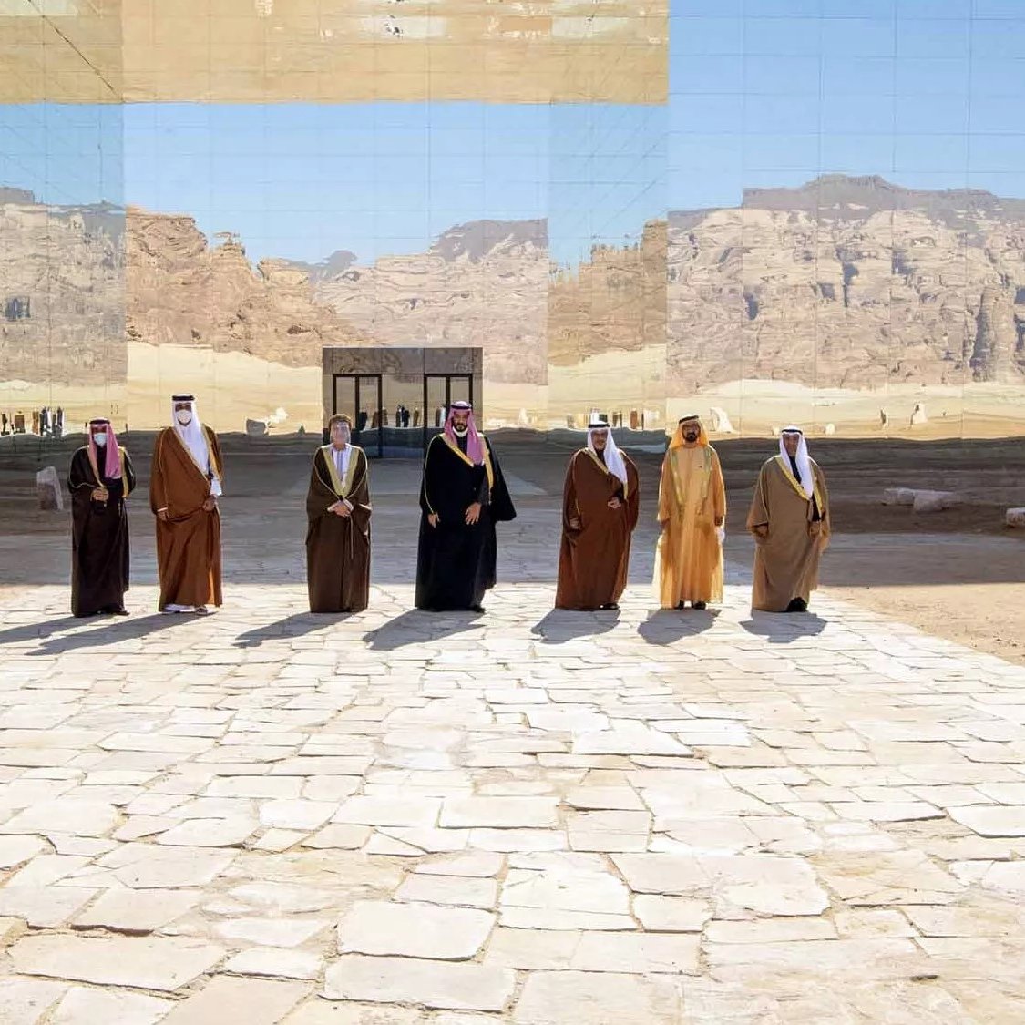 L'immagine mostra un gruppo di uomini vestiti con abiti tradizionali, dimostrando un'atmosfera di formalità e celebrazione. Sono disposti in fila su una pavimentazione di pietre chiare, con un paesaggio desertico sullo sfondo. Dietro di loro si erge una struttura riflettente che sembra integrarsi con l'ambiente circondante, creando un gioco di luci e riflessi. Le montagne in lontananza aggiungono profondità alla scena, conferendo un senso di grandiosità e atmosfera unica.