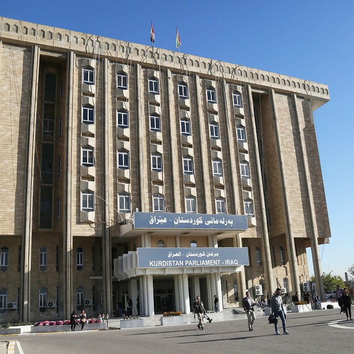 این تصویر نمای ساختمان پارلمان کردستان در عراق را نشان می‌دهد. ساختمان دارای طراحی مدرن و بزرگ است و چندین طبقه دارد. در جلوی ساختمان، پرچم‌های کردستان و عراق به اهتزاز درآمده‌اند. همچنین، افرادی در حال تردد در محوطه‌ مقابل ساختمان دیده می‌شوند. آسمان آبی و صاف به زیبایی محیط افزوده است.