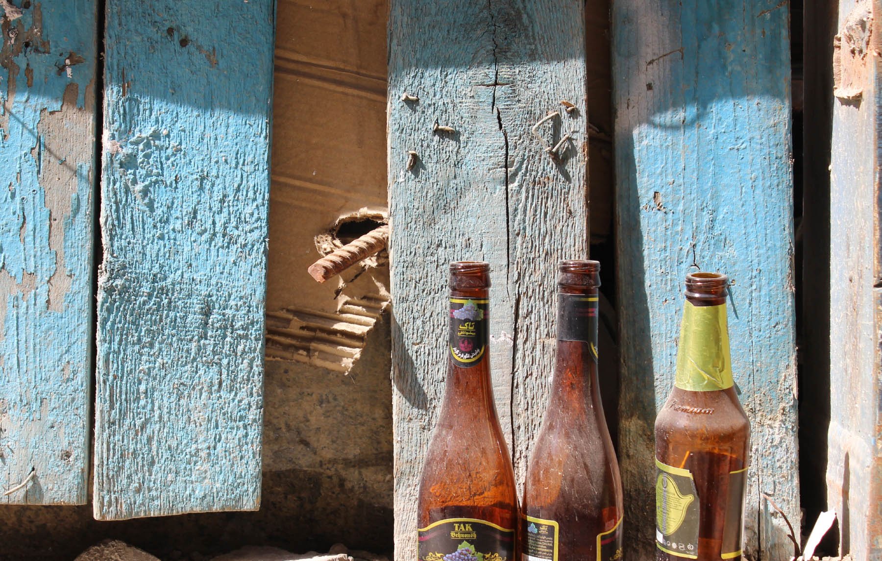 در این تصویر، سه بطری خالی در کنار دیواری که رنگ آبی دارد و کمی فرسوده به نظر می‌رسد، قرار گرفته‌اند. دیواره دارای نواحی ترک‌خورده و برخی زواید و نقص‌هاست. بطری‌ها به صورت نامرتب در کنار هم قرار دارند و بافتی کهنه و رنگ‌هایی مات دارند، که به تصویر حس قدیمی و رها شده‌ای می‌بخشد. در پس‌زمینه، تکه‌هایی از دیوار نیز قابل مشاهده است.