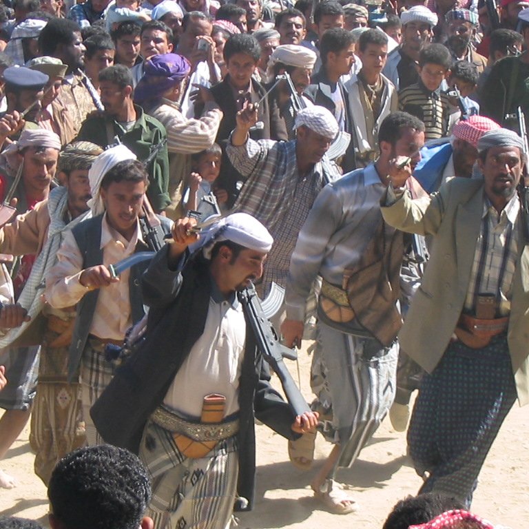 L'image montre un groupe de personnes en pleine célébration ou manifestation culturelle. Les hommes portent des vêtements traditionnels, y compris des chapeaux et des écharpes. Certains sont armés avec des fusils, ce qui pourrait indiquer un contexte de fête ou de rite traditionnel dans une région où la possession d'armes est courante. L'atmosphère semble dynamique et vivante, avec les participants se déplaçant en rythme, probablement en dansant ou en chantant.