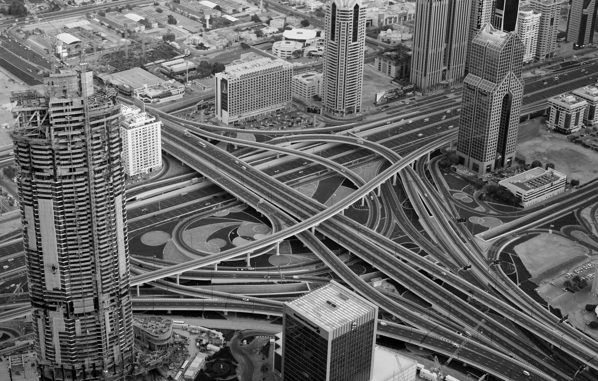 L'image présente une vue aérienne d'une métropole moderne, probablement Dubaï. On y voit de grandes tours élégantes et contemporaines, ainsi qu'un réseau complexe de routes et d'autoroutes qui s'entrelacent. Le contraste en noir et blanc accentue les formes géométriques des bâtiments et des routes, créant une impression de dynamisme et d'organisation. Des zones urbaines et dégagées suggèrent une planification urbaine sophistiquée.