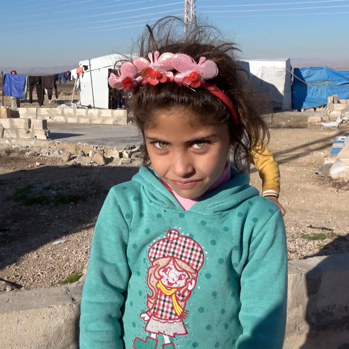 La imagen muestra a una niña pequeña en un campo de refugiados. Ella tiene largas pestañas y ojos claros, y lleva una sudadera de color azul con un diseño en la parte delantera. En su cabeza, tiene una diadema decorada con flores. El entorno es árido, con estructuras de refugio improvisadas y un cielo despejado. La expresión de la niña es curiosa y ligeramente sonriente.