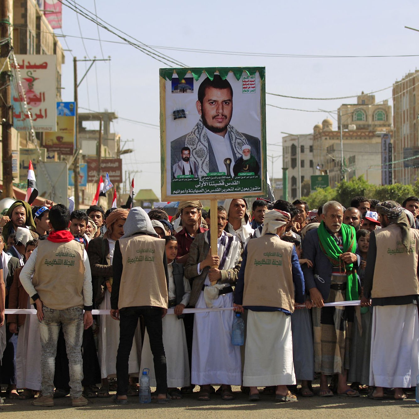 The image depicts a large crowd gathered for a rally or protest in a city, likely in Yemen. In the foreground, a group of people, some wearing beige vests, are standing behind a barricade. They are holding a large poster featuring a portrait of a man, who appears to be a political or religious leader, alongside images of other individuals. The crowd is holding flags, and buildings can be seen in the background, suggesting an urban setting. The atmosphere seems to convey a sense of solidarity or support for the figure on the poster.