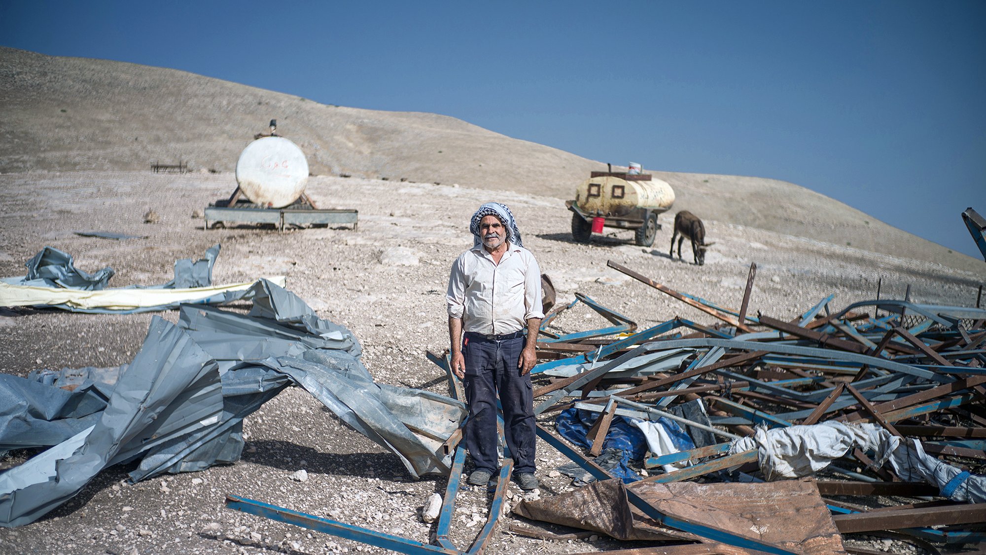 L'image montre un homme debout au milieu d'un terrain désolé. Il est entouré de débris, comprenant des structures métalliques et des morceaux de toile. En arrière-plan, on peut voir des réservoirs ou des citernes, ainsi qu'un cheval. Le paysage semble aride, avec des collines en arrière-plan sous un ciel bleu. L'homme porte une tenue simple et dégage une impression de résilience dans un environnement difficile.