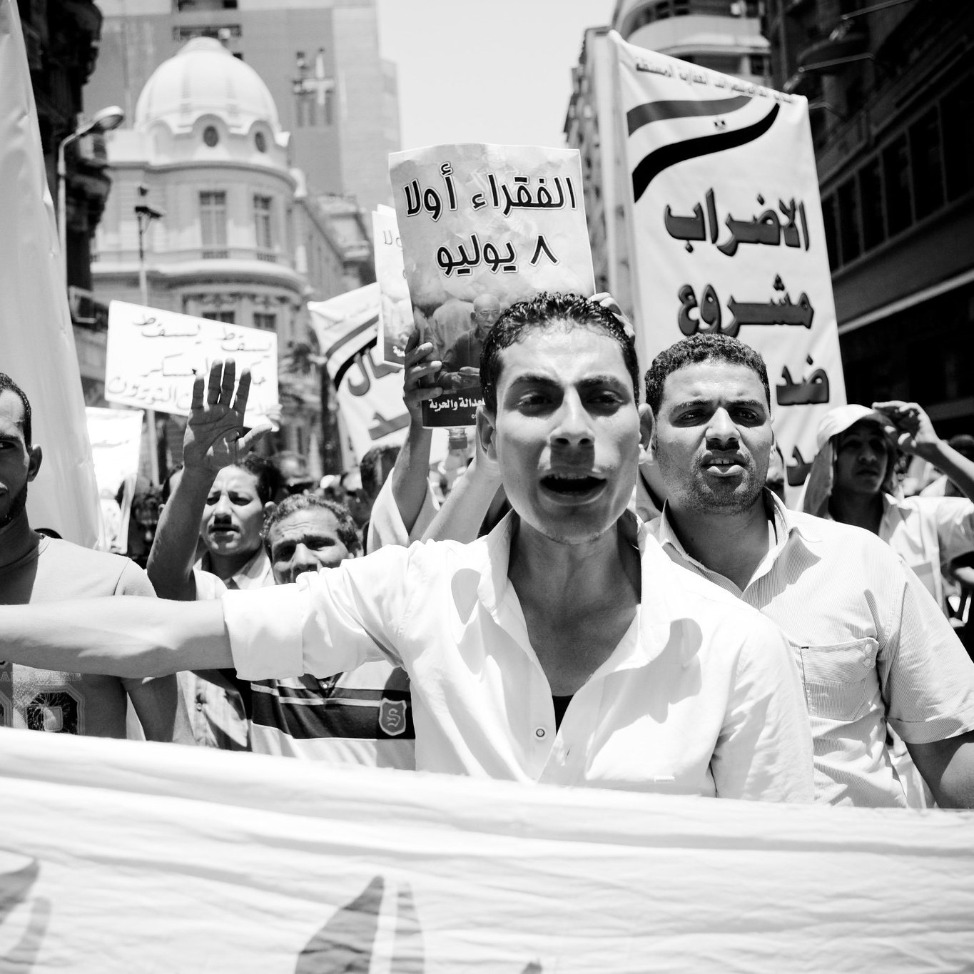 الصورة تُظهر مجموعة من المتظاهرين في شارع مزدحم، حيث يحملون لافتات ويهتفون بشعارات. يتقدم أحد المتظاهرين في المقدمة، ويبدو عليه الحماس. الجو يعكس أجواء التعبير عن المطالب والاحتجاج. تُظهر الصورة أيضاً لافتة كتب عليها "الإضراب الأول 8 يوليو"، مما يشير إلى موضوع الاحتجاج. الخلفية تحتوي على مباني حضرية، مما يضيف إلى السياق المدني للحدث.