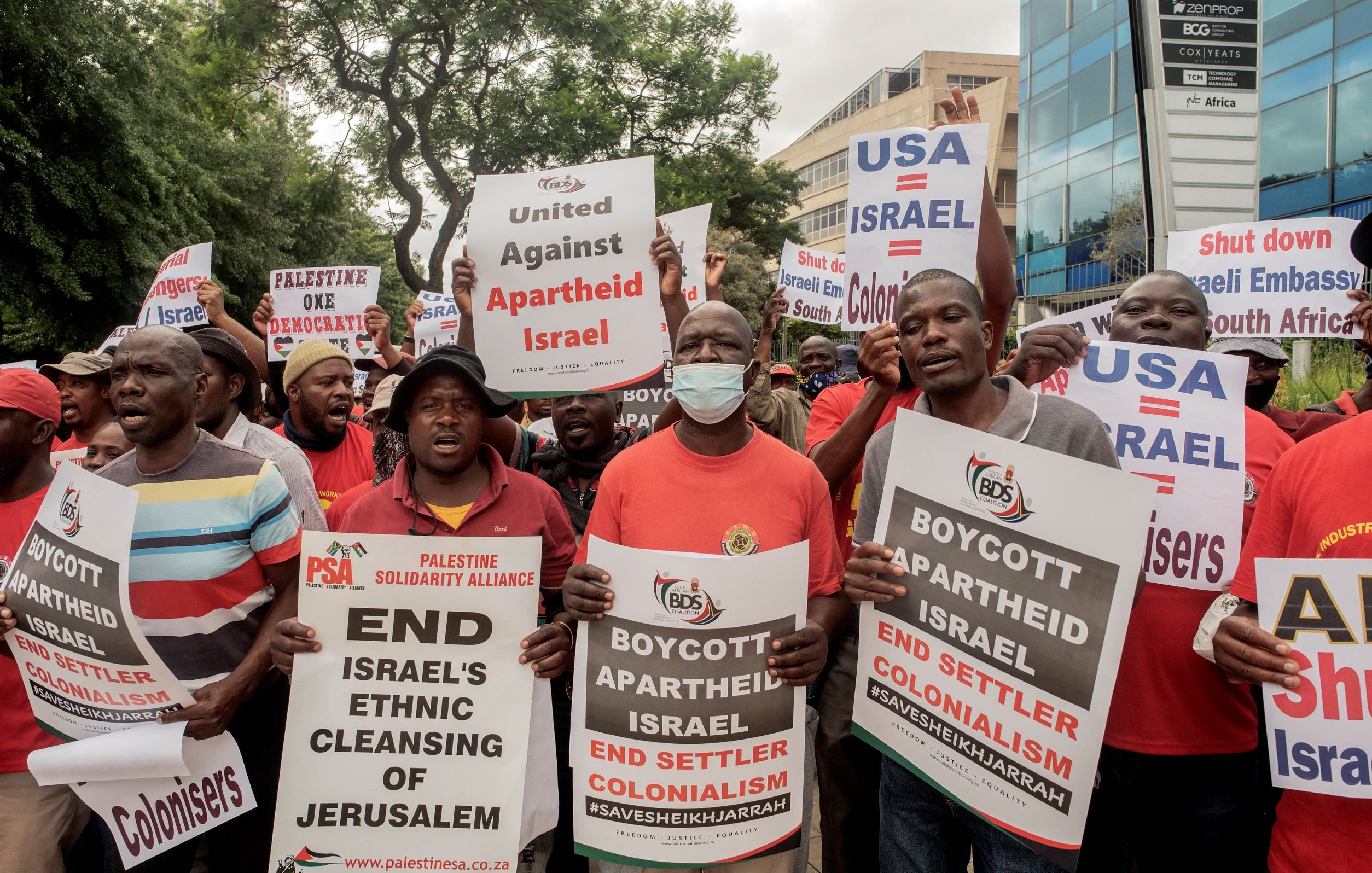 L'immagine ritrae un gruppo di manifestanti impegnati in una protesta. I partecipanti, vestiti in gran parte con magliette rosse, tengono in mano cartelli con testi che chiedono la fine dell'apartheid in Israele, il boicottaggio delle politiche israeliane e l'unità contro il colonialismo. Alcuni cartelli menzionano specificamente il "Pulizia etnica di Gerusalemme" e fanno riferimento ad azioni di protesta contro l'ambasciata degli Stati Uniti in relazione a questi temi. L'atmosfera sembra carica di energia e determinazione.