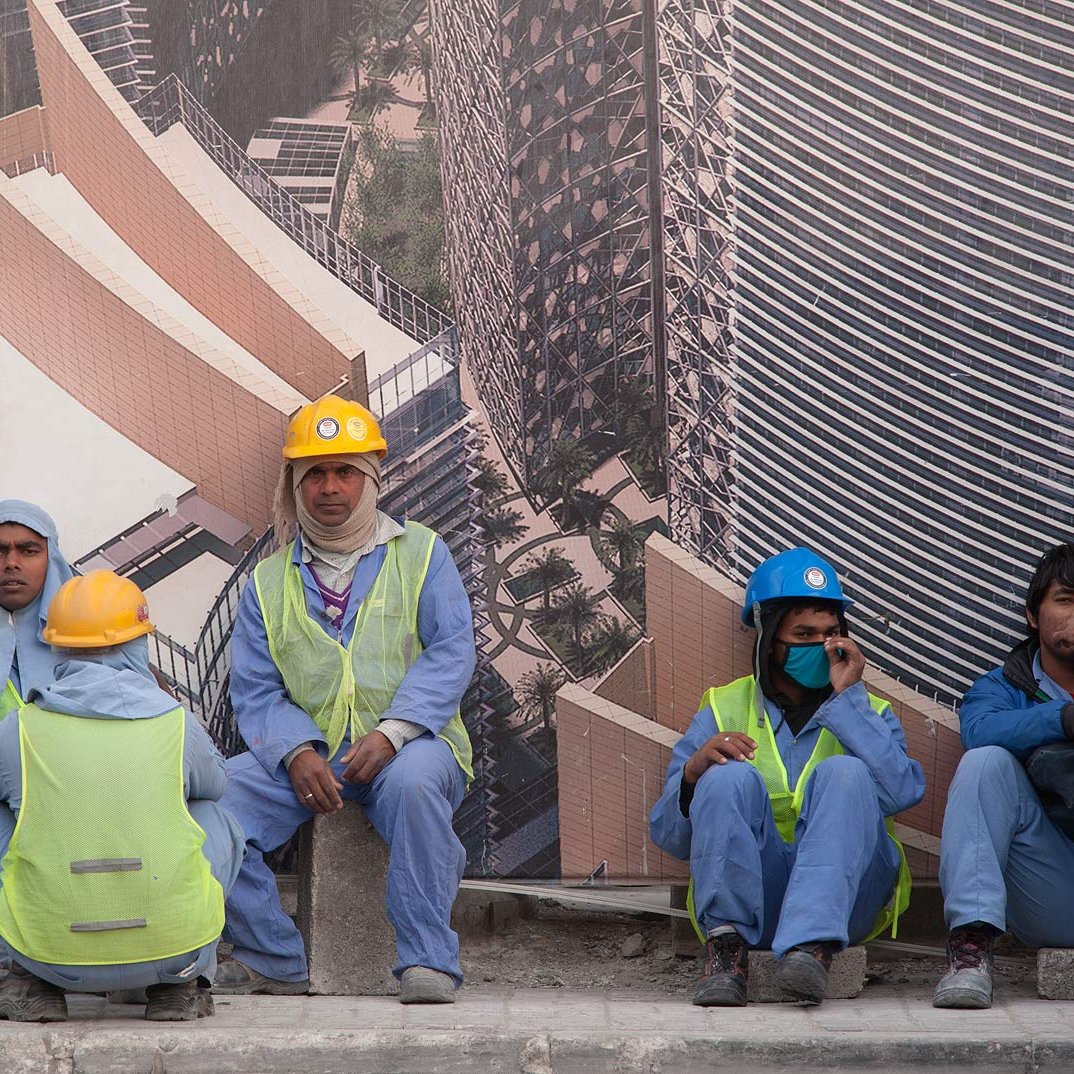En la imagen se pueden ver a varios trabajadores de la construcción sentados sobre una superficie. Están vistiendo uniformes azules y chalecos reflectantes. Algunos llevan casco de diferentes colores, como amarillo y azul. Al fondo, hay un gran cartel que muestra una imagen de un edificio moderno, sugiriendo que están trabajando en un proyecto de construcción. Los trabajadores parecen estar en un descanso, y el ambiente refleja una jornada laboral intensa.