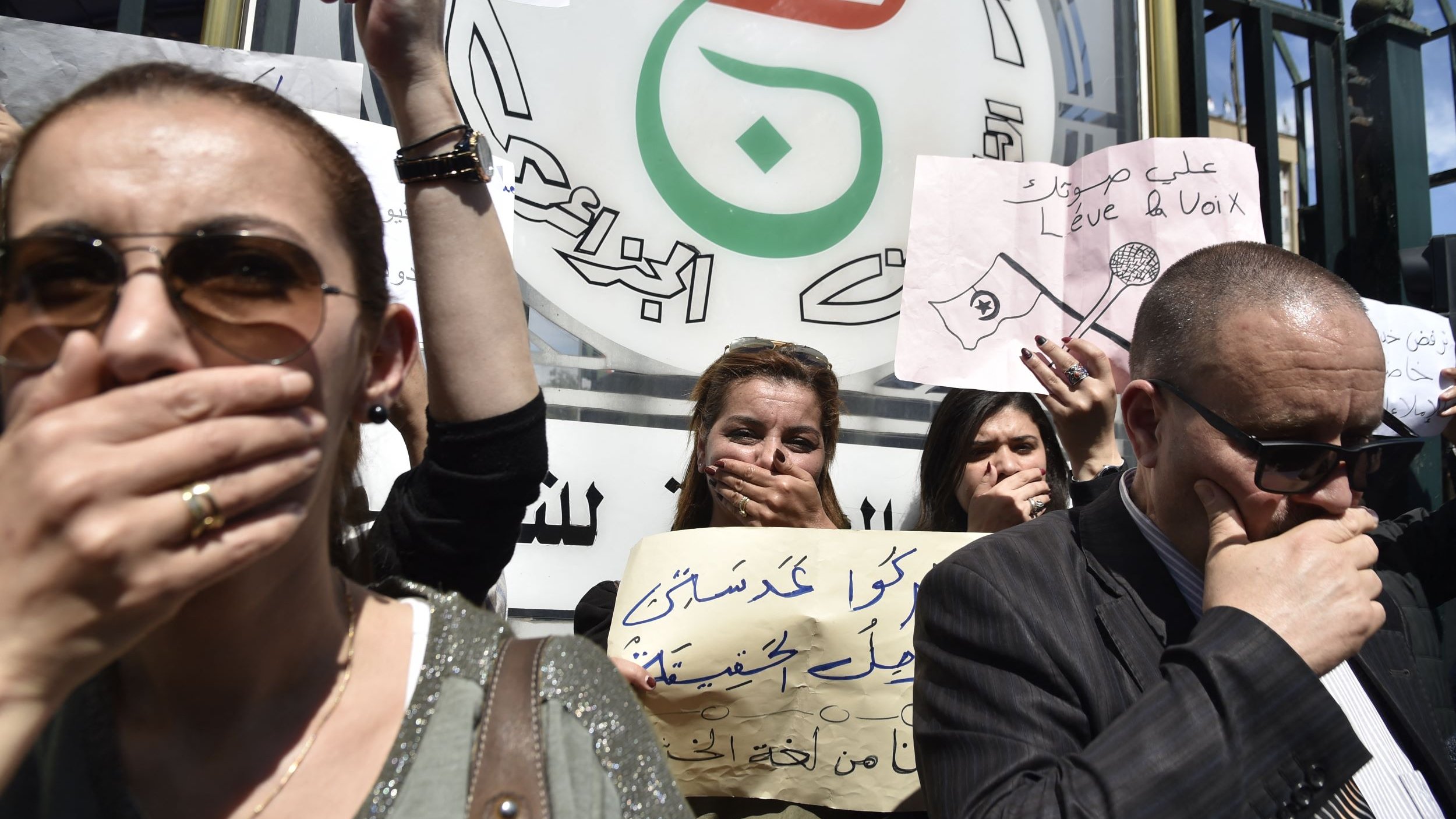 L'image montre une manifestation avec des personnes tenant des pancartes. Elles portent toutes la main sur la bouche, symbole de protestation ou de silence forcé. On peut voir des messages écrits sur les pancartes, qui semblent exprimer des revendications ou des critiques. Les manifestants sont sérieux et déterminés, reflétant un fort engagement pour leur cause. En arrière-plan, il y a un logo ou un symbole en rapport avec le mouvement.