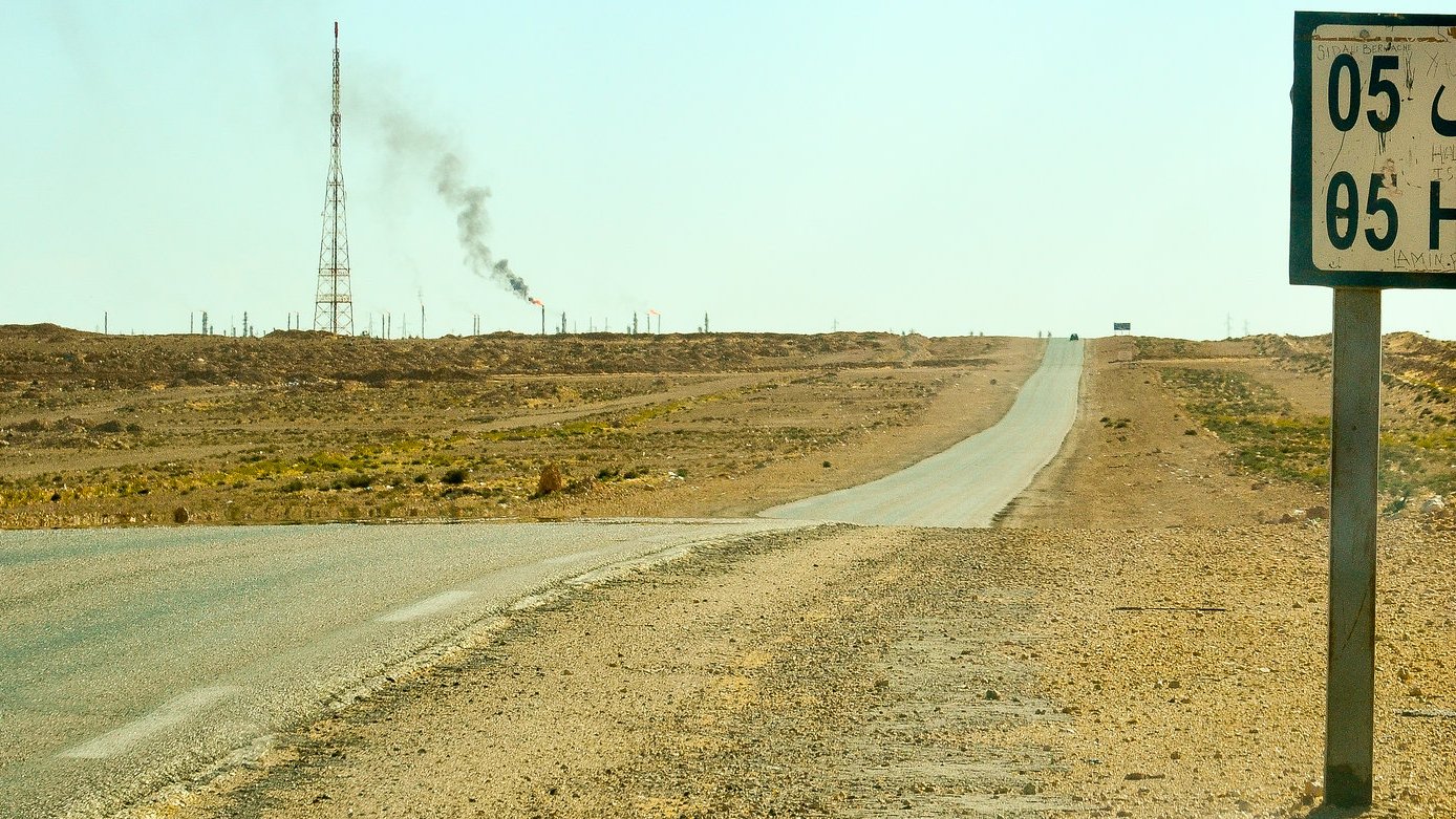 L'image montre un paysage désertique avec une route qui s'étend à perte de vue. À droite, un panneau indiquant "Hassi R'Mel" avec des chiffres. En arrière-plan, on aperçoit une tour de communication et une colonne de fumée émanant d'une installation, probablement liée à l'industrie pétrolière. Le sol est aride, et il n'y a pas beaucoup de végétation environnante, illustrant un environnement rural et isolé.