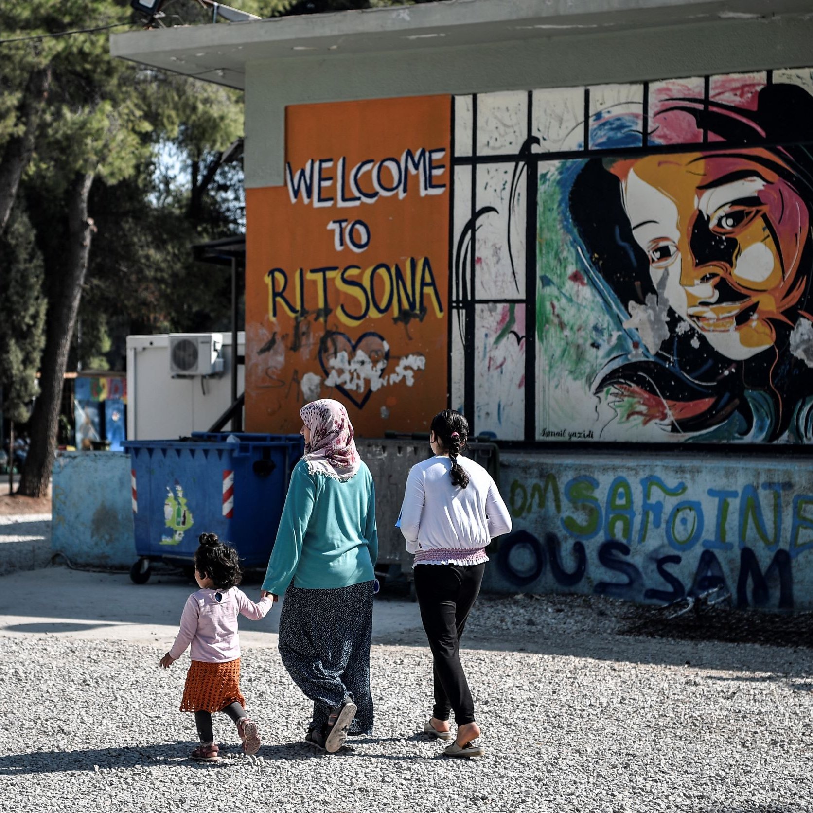 L'immagine mostra tre donne che stanno camminando in una zona all'aperto. Una di loro tiene per mano una bambina, mentre un'altra donna indossa un hijab. Sullo sfondo si può vedere un murale colorato che rappresenta un volto di donna, accompagnato dalla scritta "Welcome to Ritsona". L'area sembra essere informale e accogliente, con alberi che la circondano e strutture varie visibili.