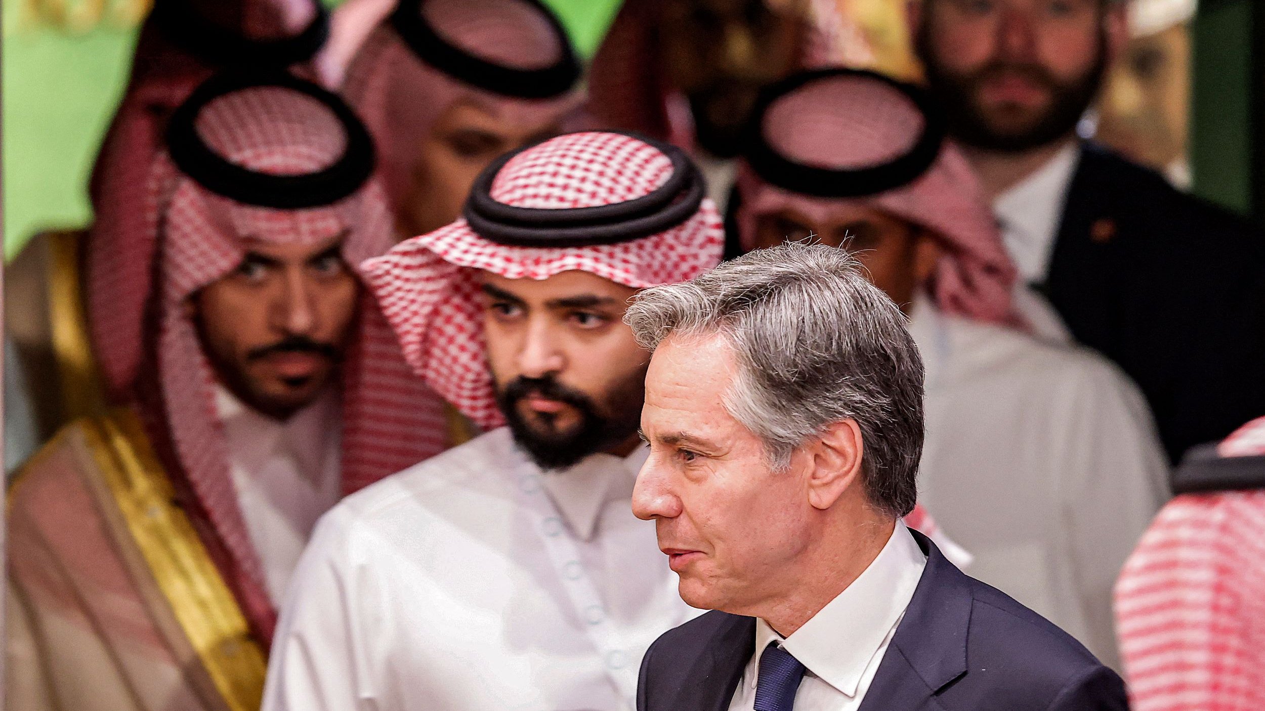 L'image montre un groupe d'hommes en vêtements traditionnels saoudiens, portant des ghoutas et des agals, en arrière-plan. Au premier plan, un homme en costume sombre marche, semblant engagé dans une conversation ou une réflexion. L'ambiance générale de l'image suggère une réunion ou un événement diplomatique.