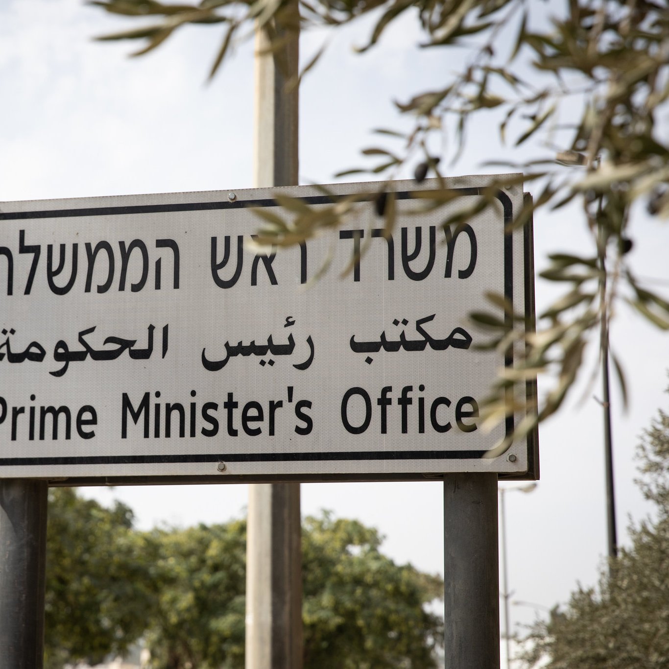 این تصویر یک تابلو راهنما را نشان می‌دهد که به زبان‌های عبری و عربی و انگلیسی نوشته شده است. متن تابلو به معنی "دفتر نخست‌وزیر" است. تابلو در یک محیط بیرونی قرار دارد و اطراف آن درختان و فضای سبز دیده می‌شود. طراحی تابلو ساده و واضح است و به نظر می‌رسد با هدف راهنمایی مردم به محل دفتر نخست‌وزیر نصب شده باشد.