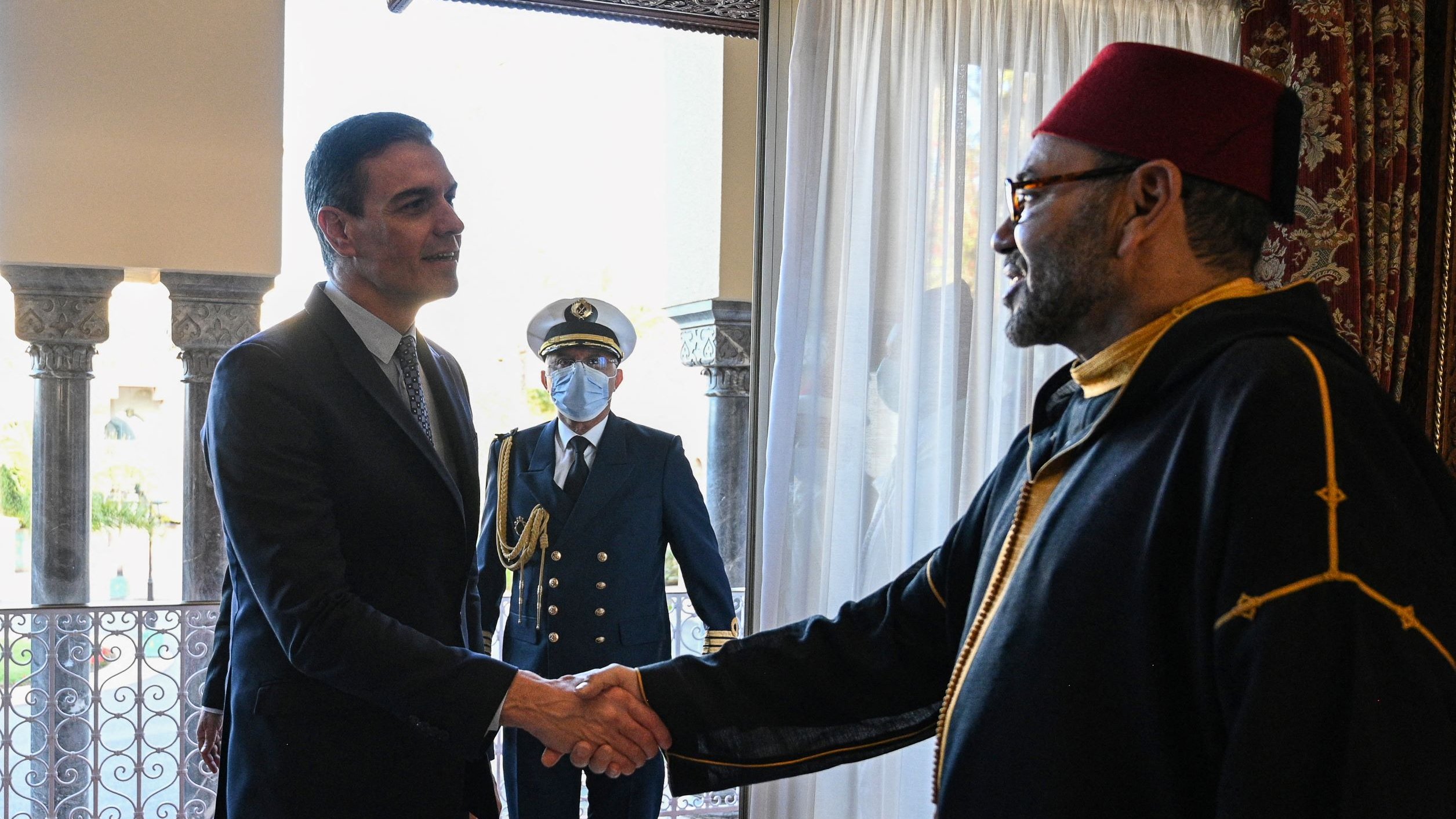 La imagen muestra a dos hombres dándose la mano en un entorno formal. Uno parece ser un líder o dignatario, vestido con un traje oscuro, mientras que el otro lleva una vestimenta tradicional, posiblemente un manto y un fez. En el fondo, se puede ver a un tercero, que es un guardia o oficial en uniforme, observando la escena. La decoración del lugar es elegante, con cortinas blancas y un entorno arquitectónico notable.