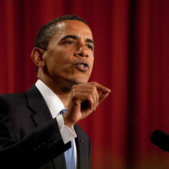 L'image montre un homme en costume noir avec une chemise blanche et une cravate bleue. Il est en train de parler avec passion, levant un doigt pour souligner un point important. En arrière-plan, on peut voir un drapeau ou un décor formel. L'atmosphère semble très sérieuse et engageante, suggérant qu'il est en train de prononcer un discours ou de faire une déclaration importante.