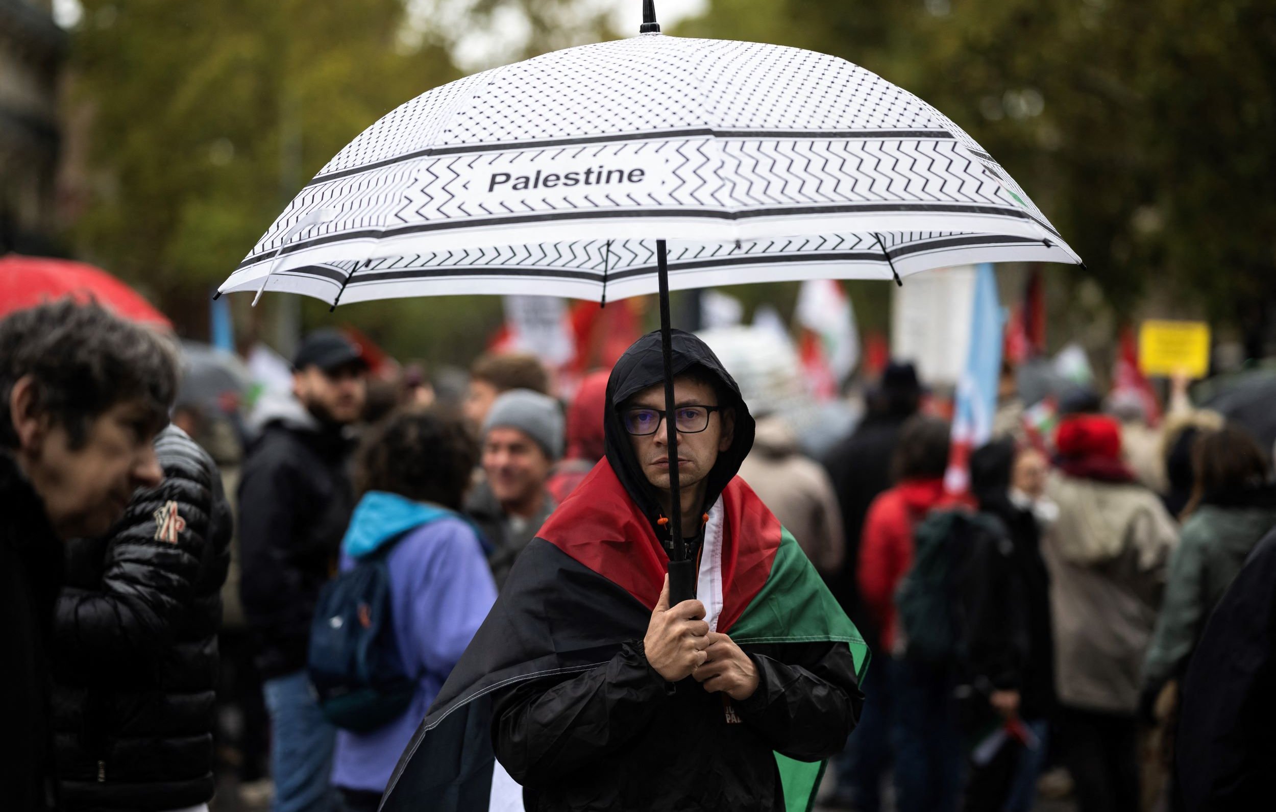 The image depicts a gathering, likely a protest or demonstration, where a person stands under a black-and-white umbrella labeled "Palestine." This individual is draped in a Palestinian flag and wears glasses, displaying a serious expression. Surrounding them are other participants, some holding flags and signs, with a backdrop of trees and a cloudy sky, suggesting an outdoor setting and possibly inclement weather. The scene captures a moment of solidarity and advocacy related to Palestinian issues.