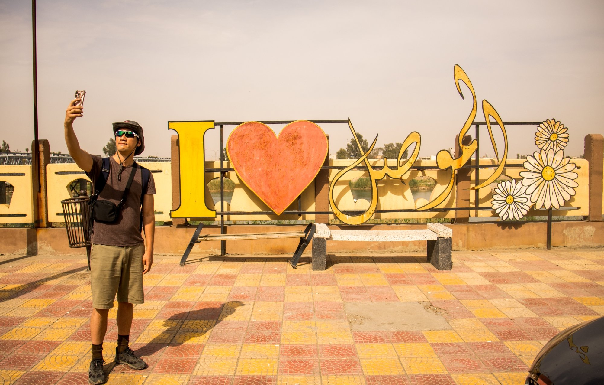 Un uomo fa un selfie davanti a un grande cartello colorato con "I ❤️" e fiori.