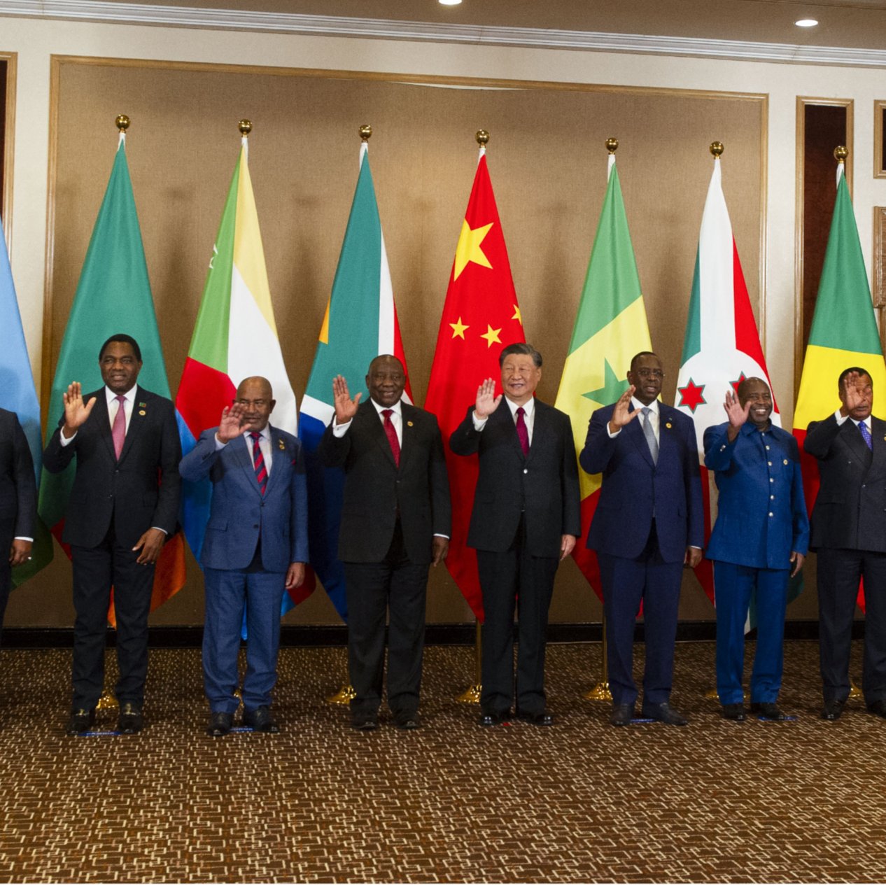 La imagen muestra a un grupo de líderes y representantes de diferentes países alineados en una sala. Detrás de ellos hay banderas de varias naciones, incluidos países africanos y de otras partes del mundo. Todos los individuos parecen estar posando para una fotografía, algunos levantando la mano en señal de saludo. La decoración del lugar es elegante, con un ambiente formal que refleja la importancia del evento.