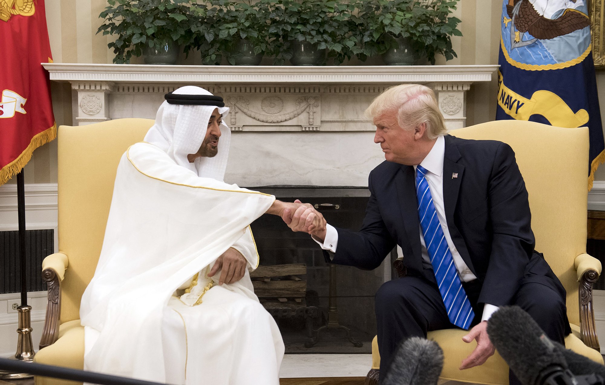 En la imagen se puede ver a dos hombres en un ambiente formal. Uno de ellos viste una túnica blanca y una kefía, que son vestimentas tradicionales de algunos países árabes, mientras que el otro lleva un traje oscuro con una camisa blanca y una corbata azul. Ambos están en un apretón de manos, lo que sugiere un acuerdo o una conversación amistosa. Al fondo, se aprecia una decoración sobria y plantas, así como banderas que indican que se encuentran en un lugar oficial, probablemente una oficina o sala de reuniones.