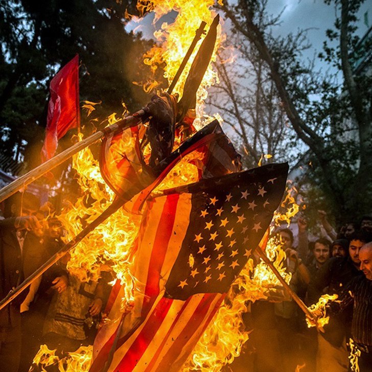 L'image montre une scène de protestation où des gens brûlent un drapeau américain. Les participants, visiblement passionnés, lèvent les bras et brandissent des bâtons. L'atmosphère est intense, avec des flammes qui s'élèvent autour du drapeau, tandis que des arbres et des bâtiments en arrière-plan ajoutent à la dynamique de la situation. Cette action symbolise souvent une forte opposition ou un message politique.