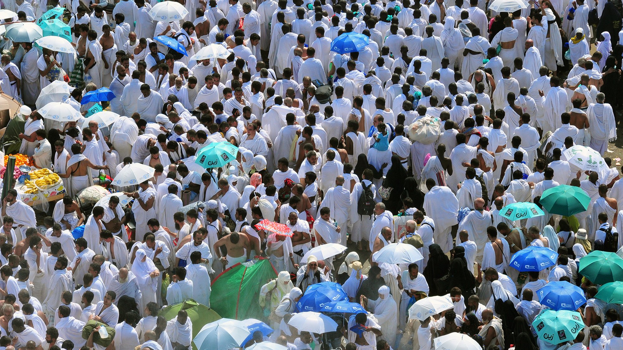 L'image montre une grande foule de personnes vêtues de vêtements blancs, probablement en train de participer à un pèlerinage ou à un événement religieux. De nombreux participants arborent des parapluies colorés, qui offrent de l'ombre dans un environnement ensoleillé. L'atmosphère semble très animée, avec un grand nombre de personnes rassemblées, ce qui reflète un moment de rassemblement spirituel.