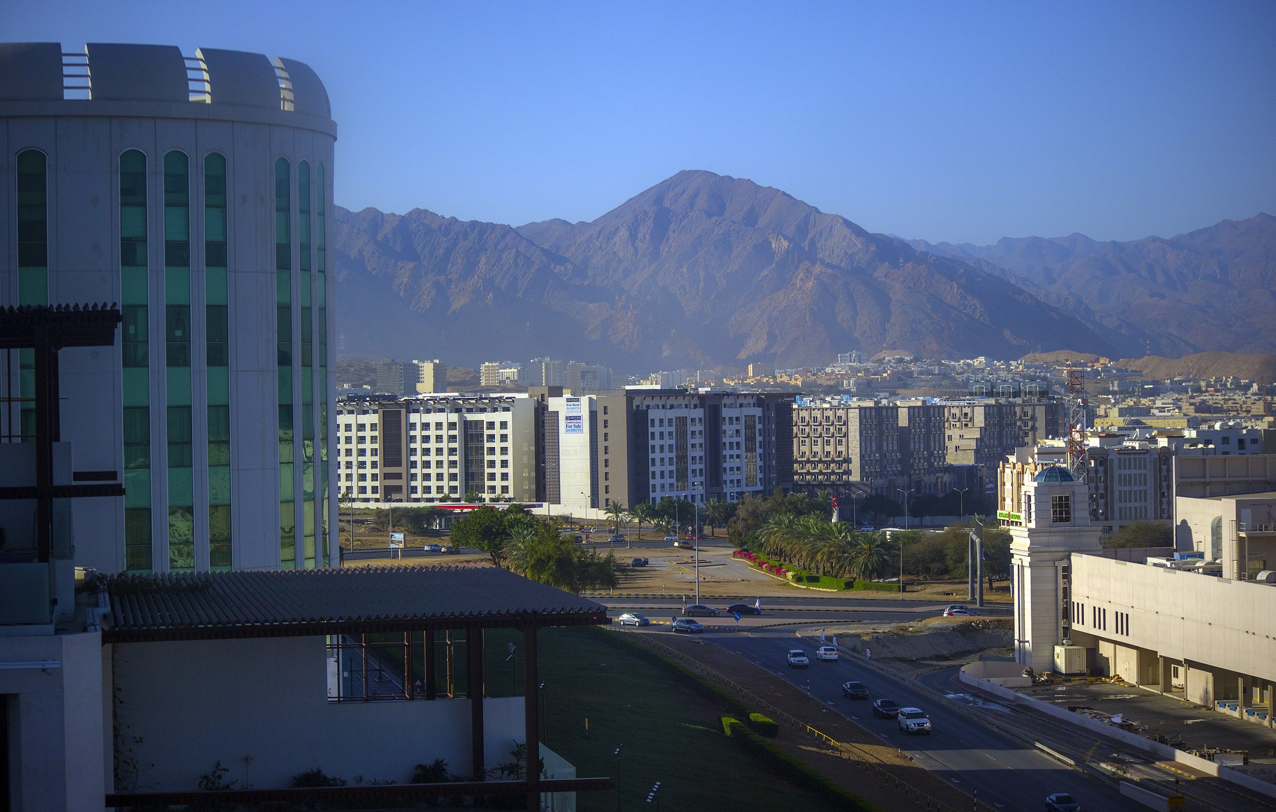 La imagen muestra un paisaje urbano con una mezcla de edificios modernos y montañas al fondo. A la izquierda, se puede ver un edificio de gran altura con un diseño contemporáneo. En el centro, hay una serie de edificios de varios pisos, que parecen ser parte de un área urbana. Al fondo, se aprecian montañas con una forma escarpada y un color terroso, indicando un ambiente montañoso. La luz del sol ilumina la escena, generando un ambiente claro y fresco. En primer plano, hay una carretera que conecta los diferentes sectores de la ciudad.