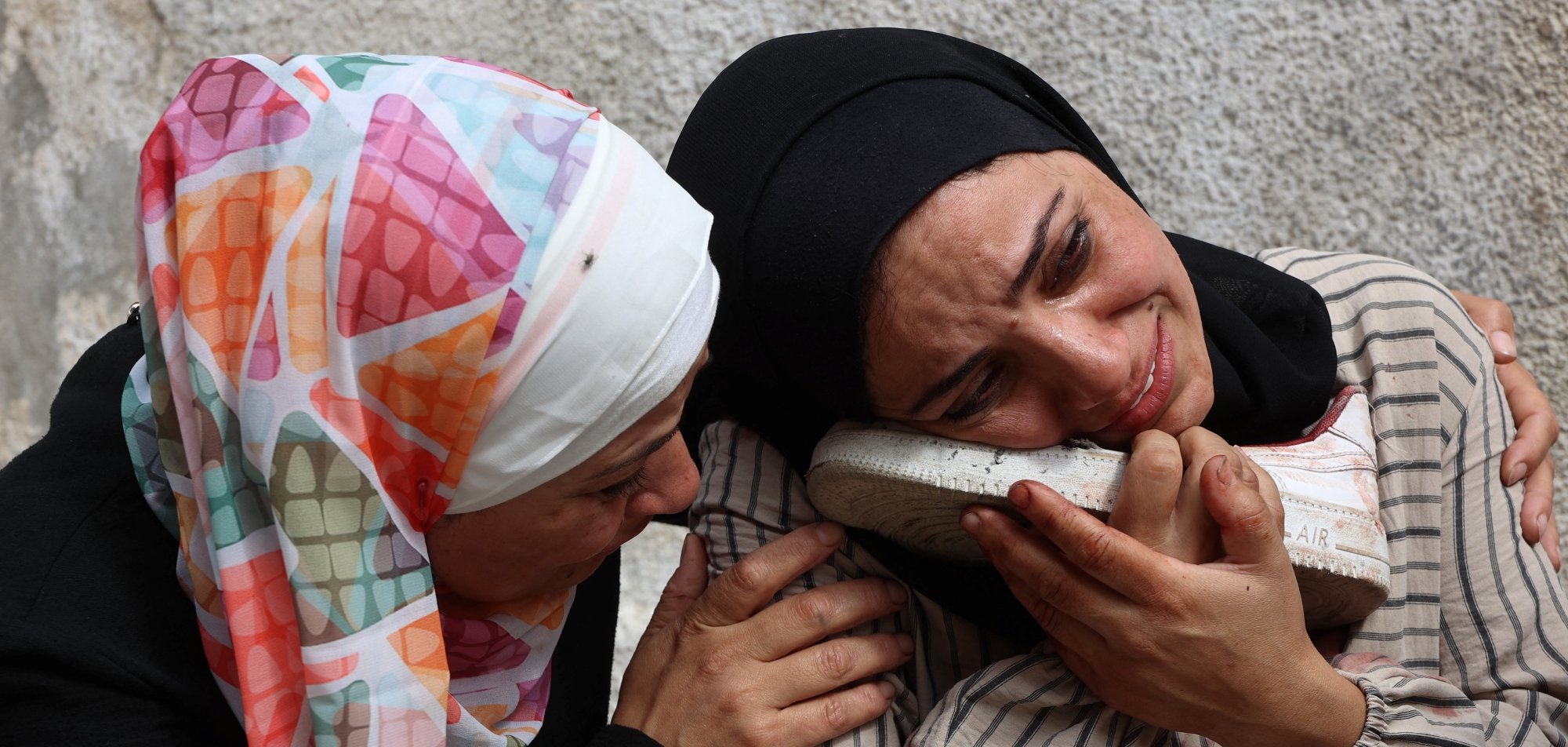 Two women in headscarves are embracing, expressing deep sorrow, with one holding a shoe.