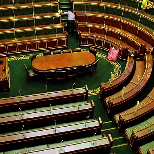 تظهر الصورة قاعة اجتماعات برلمانية، حيث تتوزع المقاعد بشكل دائري حول طاولة مستطيلة في المنتصف. المقاعد مصممة بطريقة تتسع لعدد كبير من الأعضاء، بينما يبدو الجو هادئًا. بعض العمال يقومون بتنظيف القاعة، مما يوحي بأن الاجتماع قد انتهى أو لم يبدأ بعد. الأرضية مزينة بنمط معين، مما يضيف جمالية للمكان.