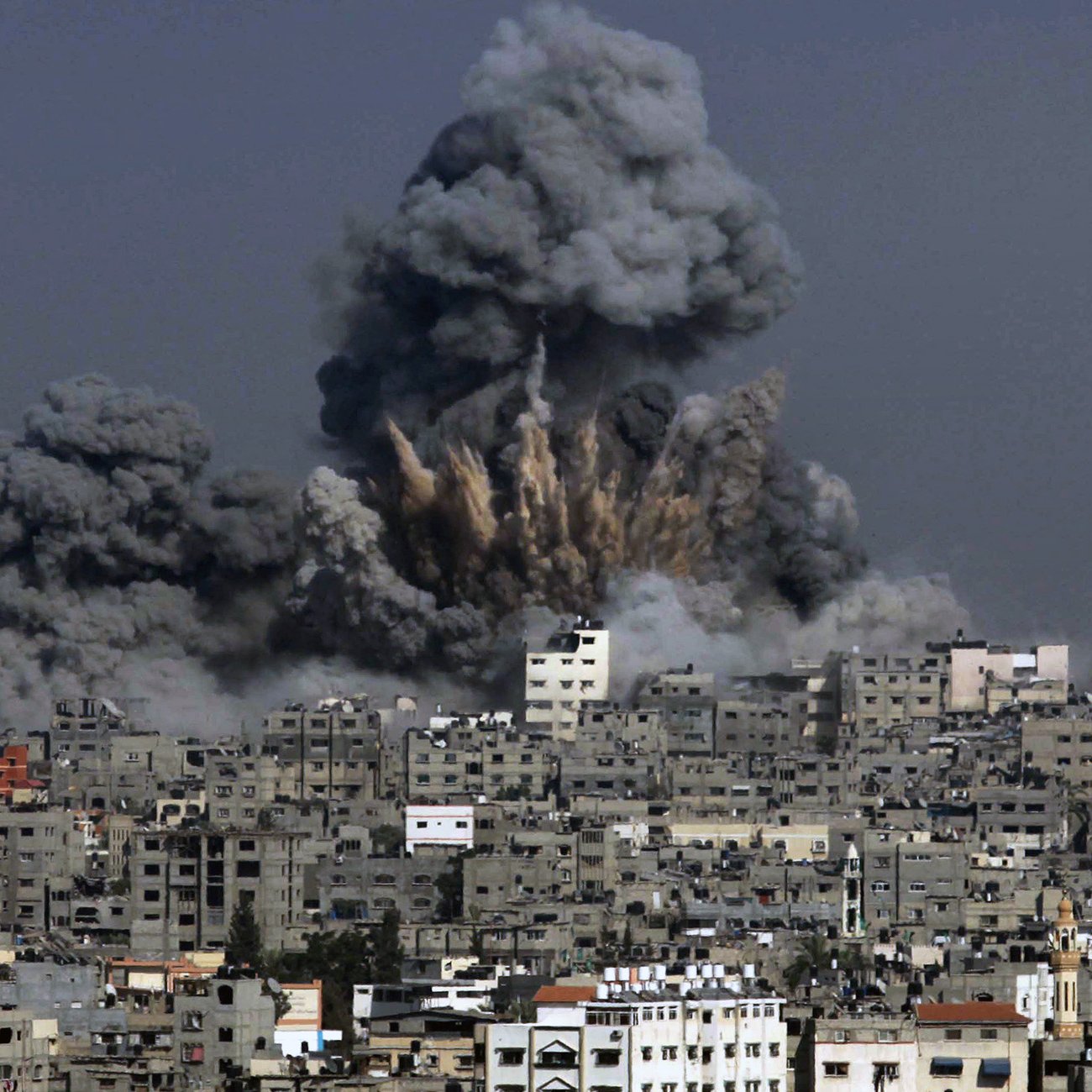 The image depicts a scene of destruction with a large explosion occurring in an urban area. Thick, dark gray smoke billows into the sky, rising above the buildings in the foreground. The skyline is dominated by closely packed structures, showing signs of damage. The overall atmosphere conveys a sense of chaos and devastation, characteristic of an area affected by conflict or military action.